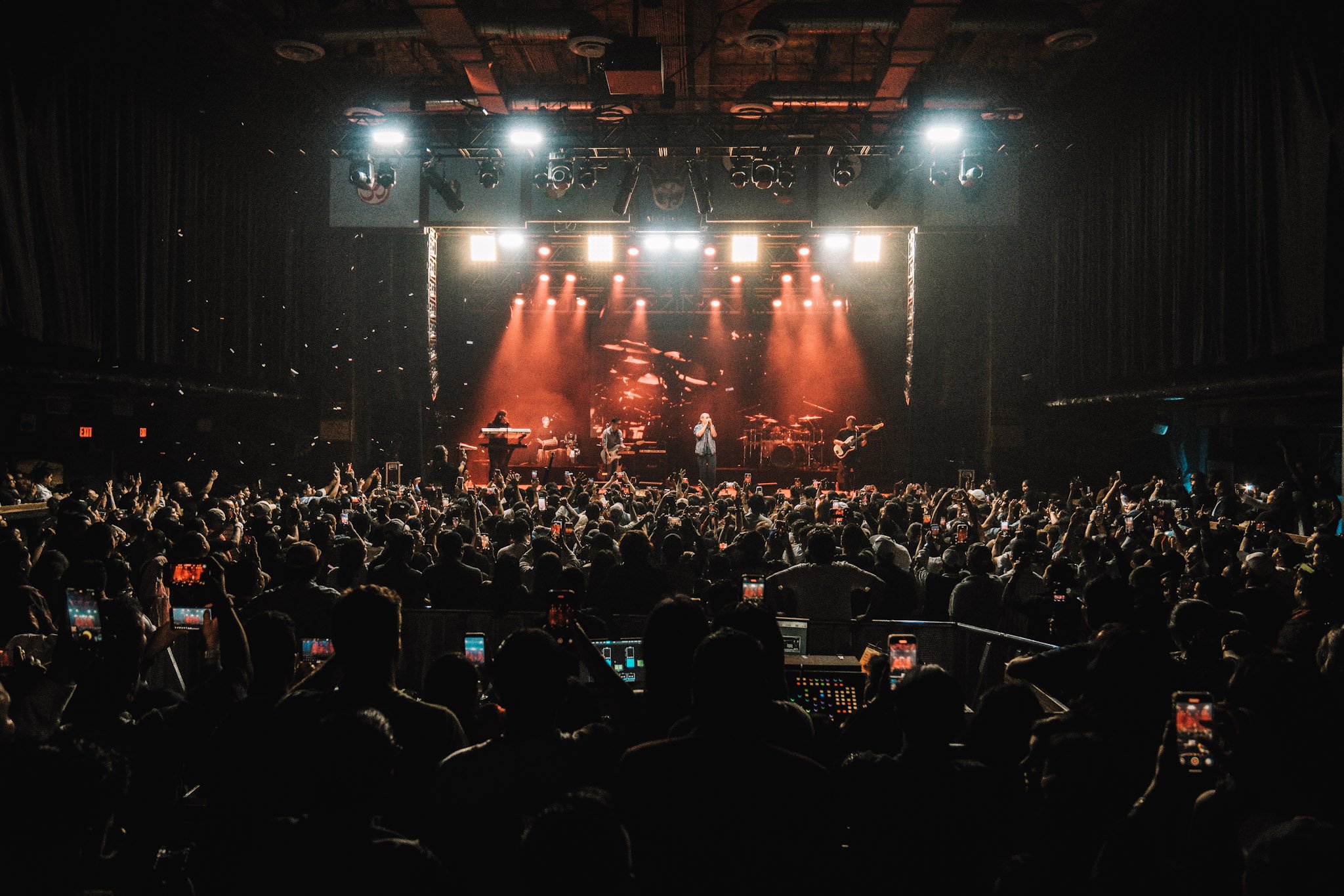 A live concert with a band performing on stage surrounded by lighting effects, while the audience in the dark attends and records the performance on their phones.