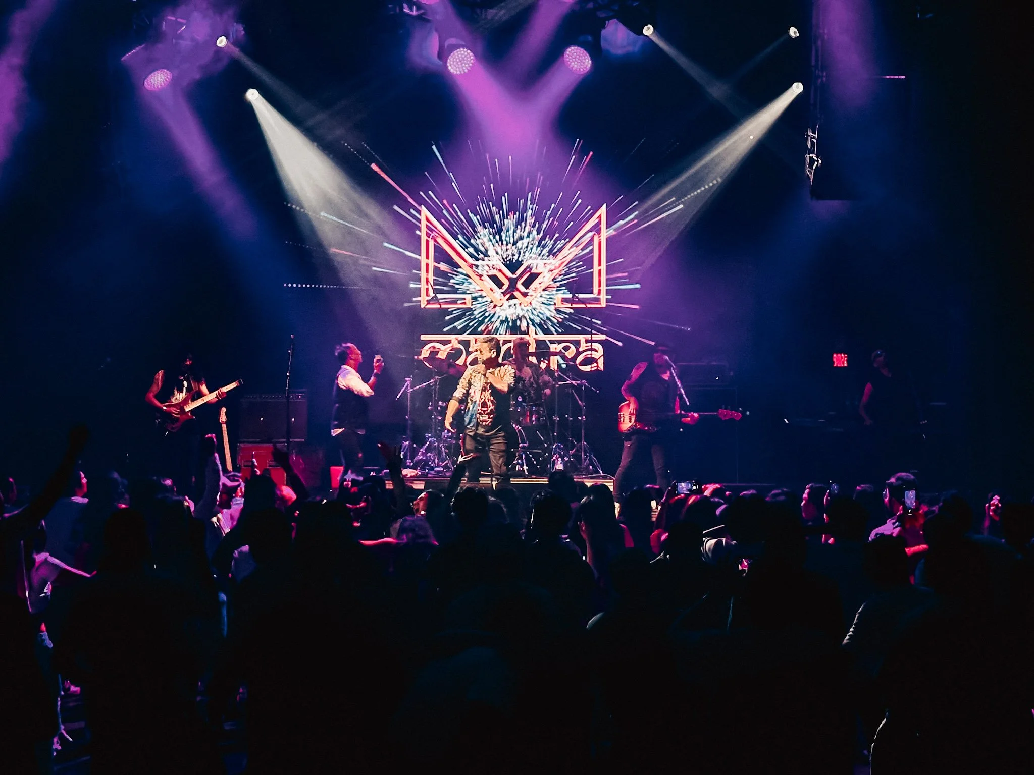 A live concert with a band performing on stage, colorful lights, and an audience watching.