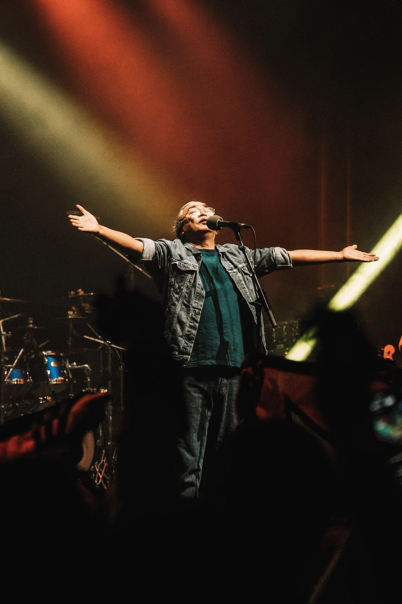 A man on stage with arms outstretched, singing into a microphone during a concert, with colorful stage lighting and drums in the background.
