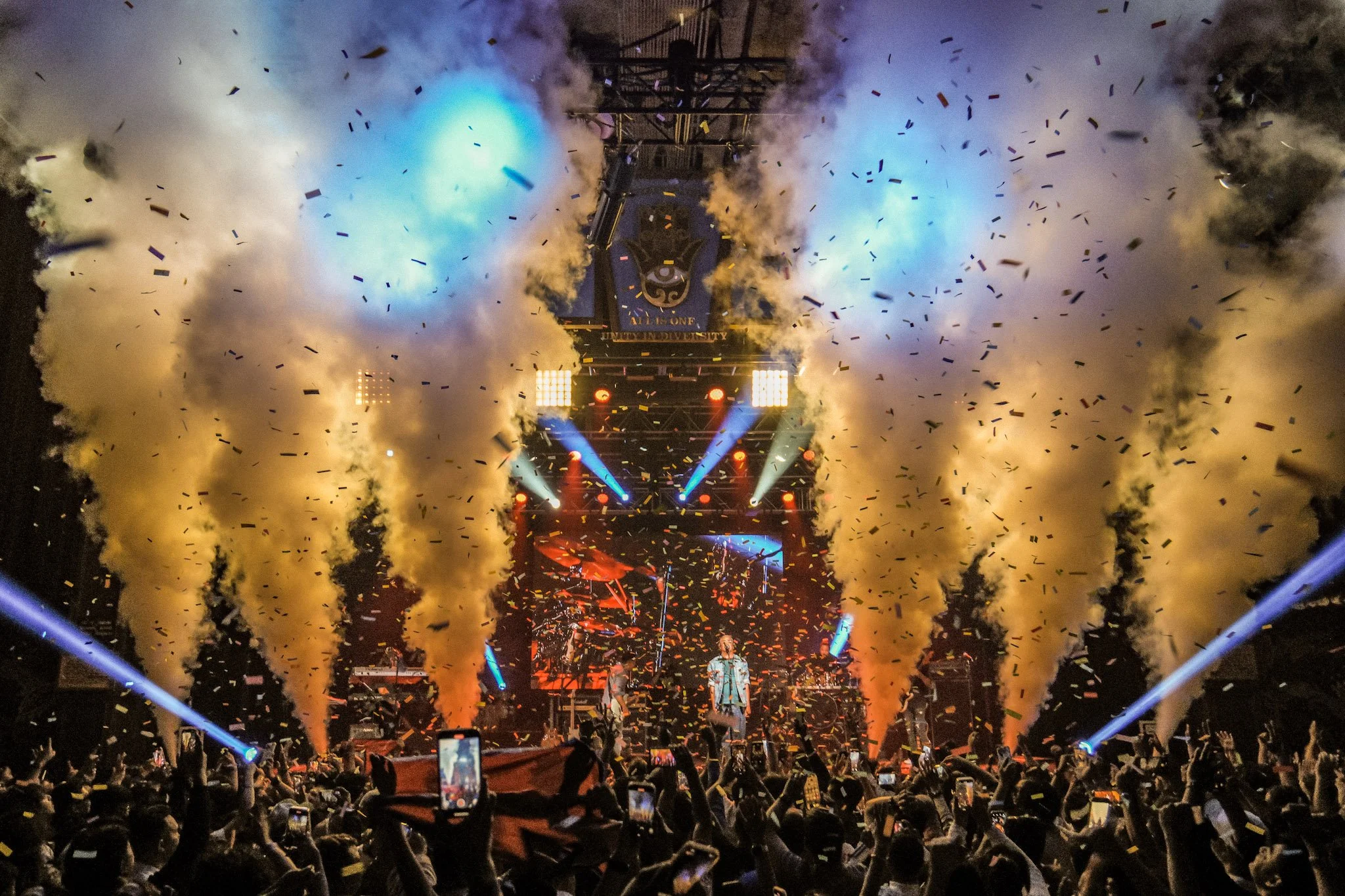 A large concert stage with performers, dramatic lighting, fog, and confetti falling. Audience members are capturing the moment with their phones.