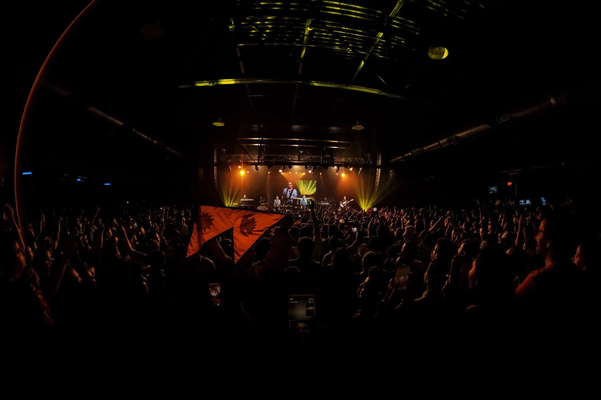Crowd at a concert or music festival with a person holding a Canadian flag in front. Stage with musicians and colorful lights in the background.