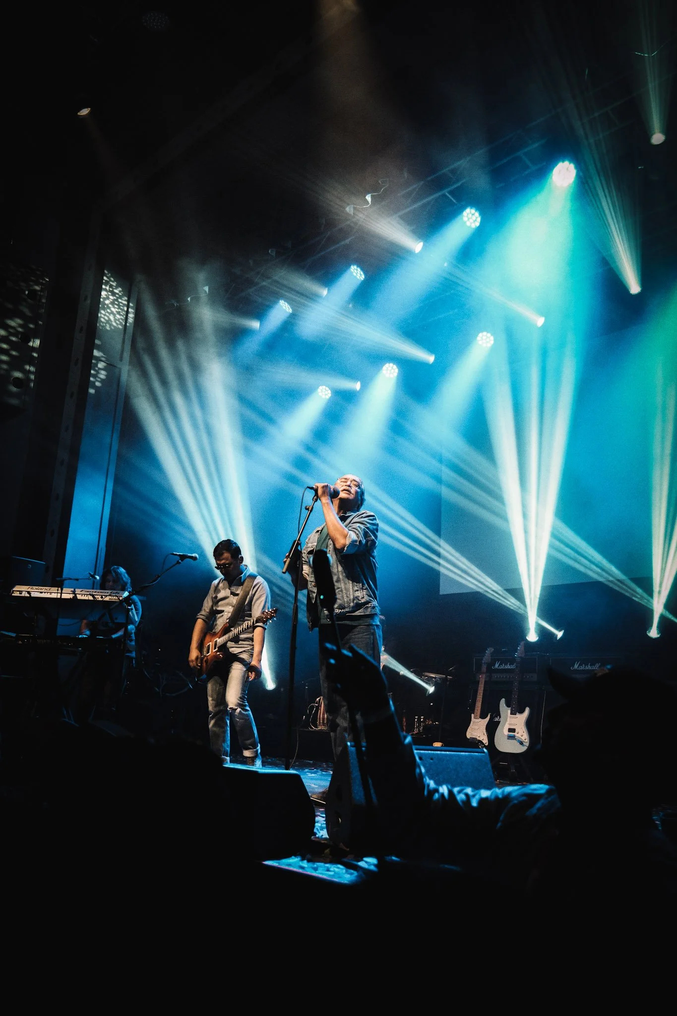 A live music performance on stage with three musicians. The lead singer is holding a microphone with lights shining behind him. Two other musicians are playing guitars and keyboard, with colorful stage lighting in the background.