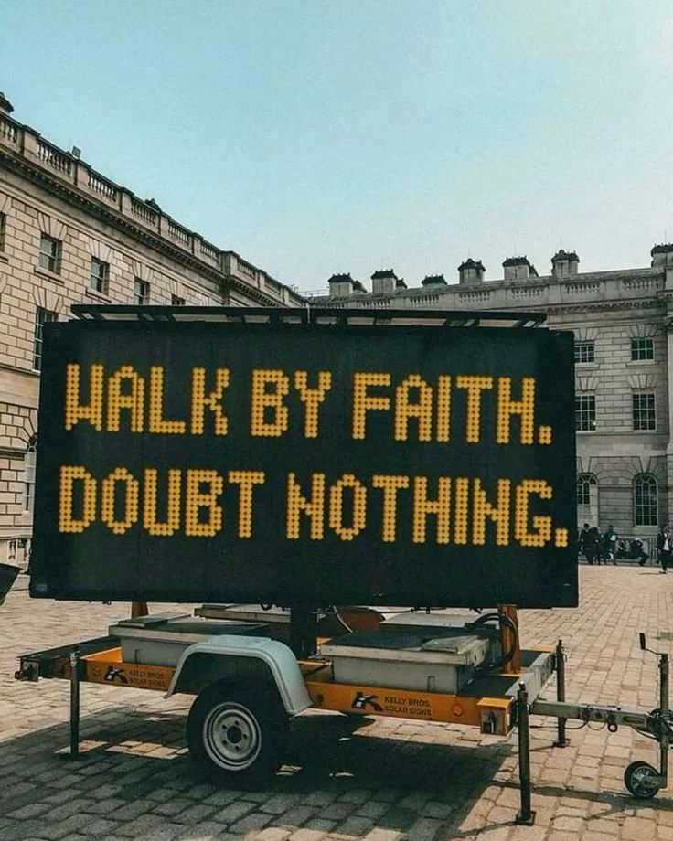 A mobile digital sign with yellow letters on a black background displaying the message "Walk by faith. Doubt nothing." in an urban setting with historic buildings.