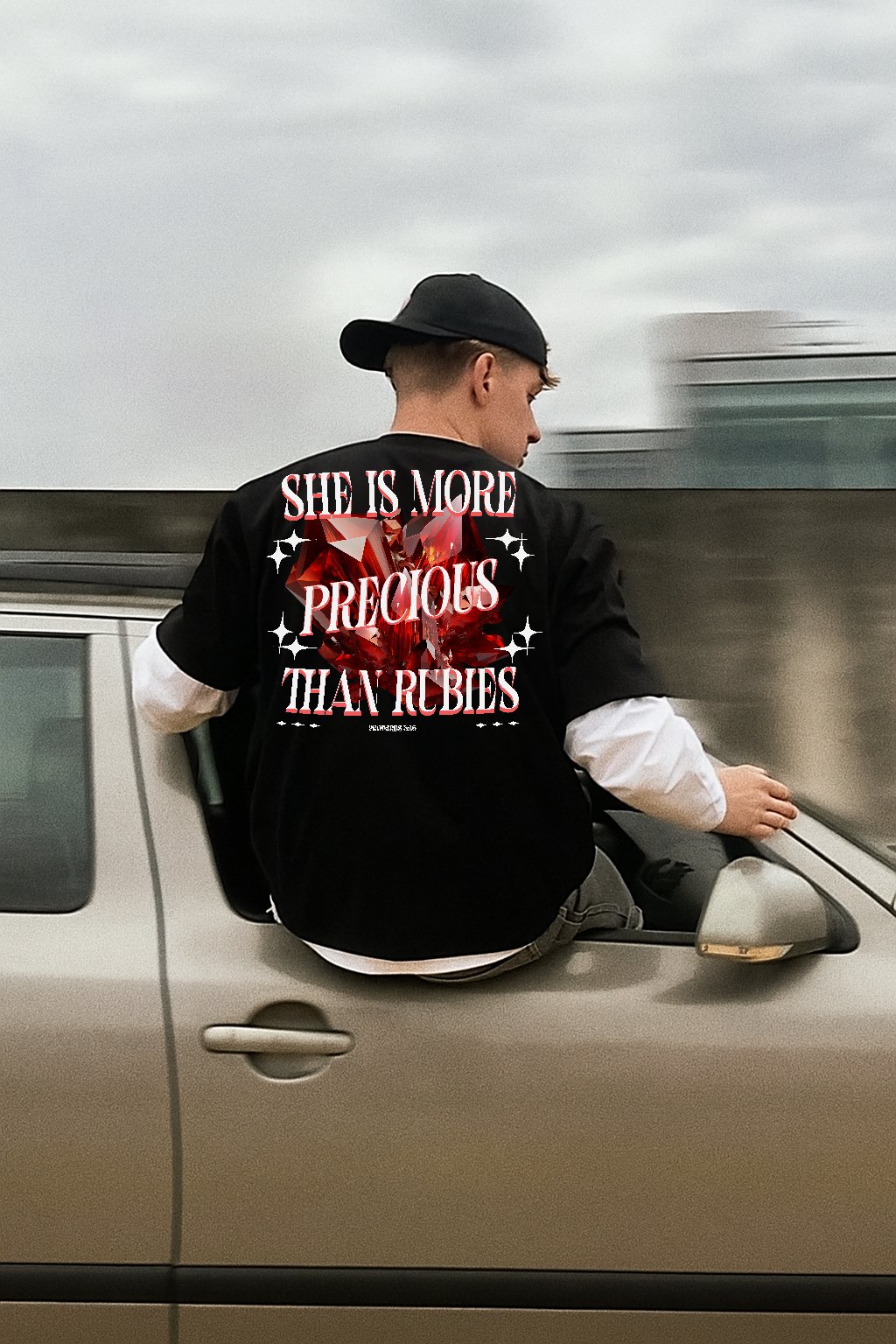 A young man with a black cap and black shirt with white sleeves sitting on the hood of a silver car, looking to the side, with a cloudy sky in the background. The back of his shirt has a graphic of a red gemstone with the text