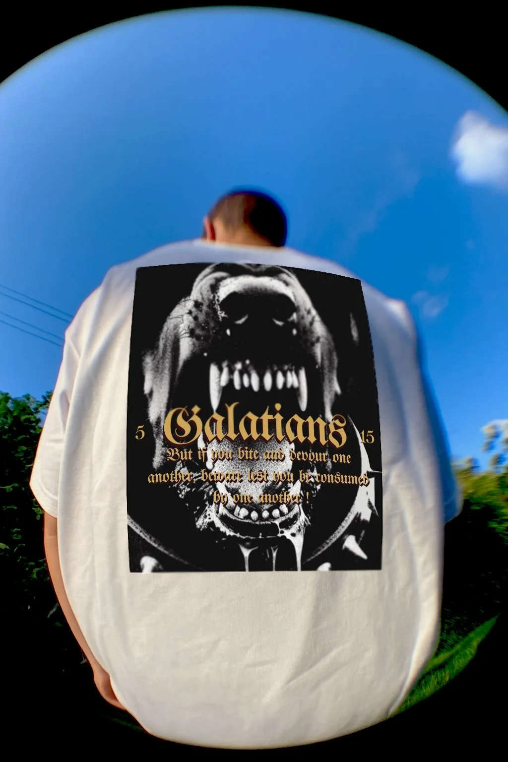 Person wearing a white T-shirt with a large black and white graphic of a fierce dog and the words '5 Galatians 5:15 But if you bite and devour one another, beware lest you be consumed by one another!' printed on the back, outdoors with a blue sky and some greenery in the background.