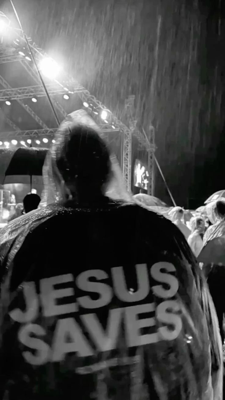 A person at an outdoor event during rain, wearing a jacket that says 'Jesus Saves' in large letters, with a stage and crowd visible in the background.