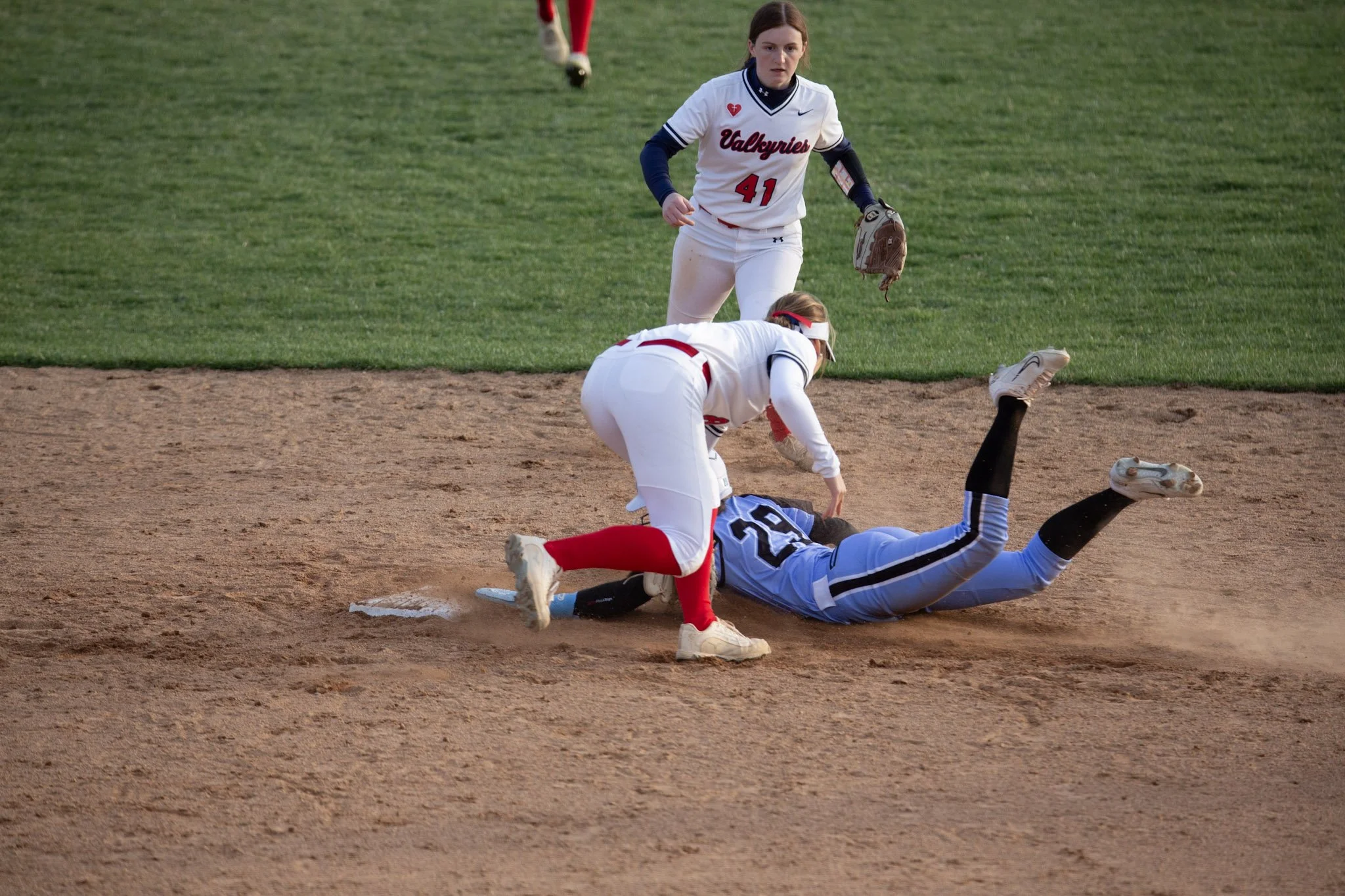 Two softball players in white uniforms with red and blue accents on a dirt field, one falling on the ground and the other reaching down to assist, with a third player standing nearby.