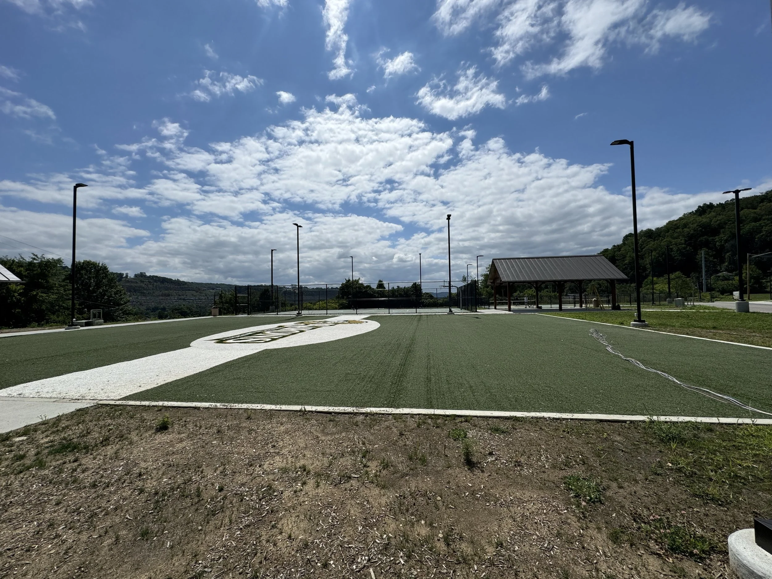 An outdoor sports court with artificial green turf, surrounded by several tall light poles, a small shelter with a metal roof, and hills with trees in the background. The sky is partly cloudy with scattered clouds.