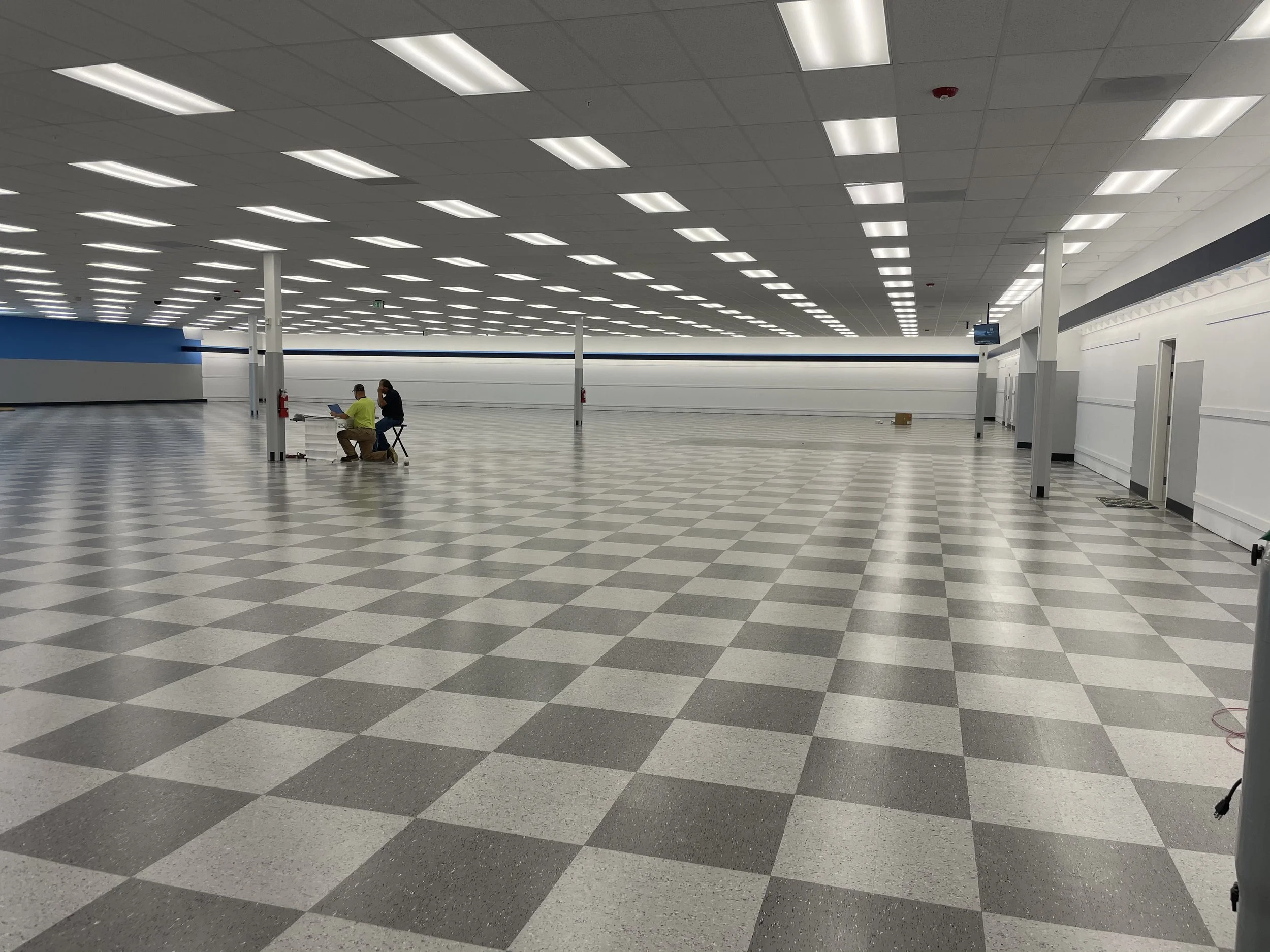 Empty airport terminal with checkered gray and white floor, white ceiling with fluorescent lights, white and blue walls, and two people sitting on chairs working on laptops.