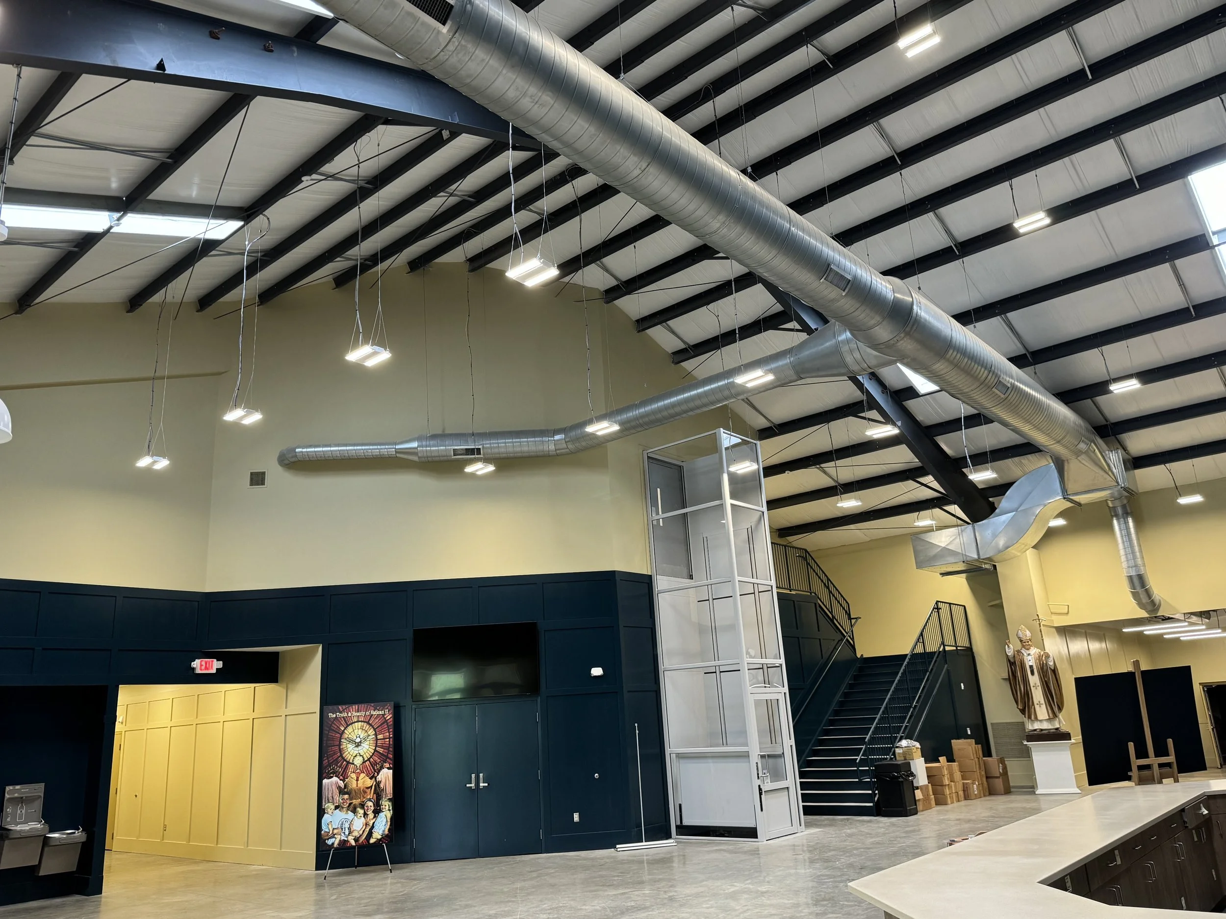 Interior of a modern building with a high ceiling, exposed metal ductwork, stairs, and religious statues.