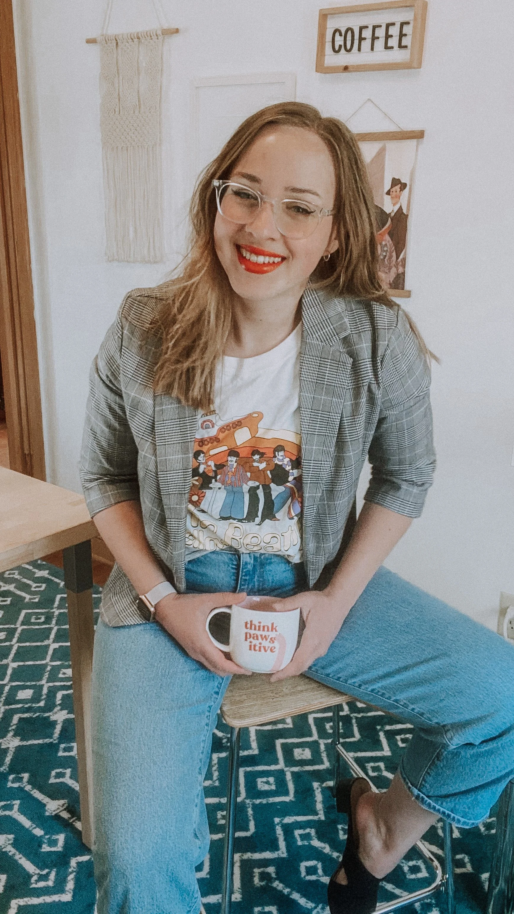 Woman sitting on a wooden chair holding a white mug with the words 'think pawsitive' in red, wearing glasses, a graphic t-shirt, a plaid blazer, and blue jeans, in a cozy room with wall art including a 'COFFEE' sign and a tapestry.