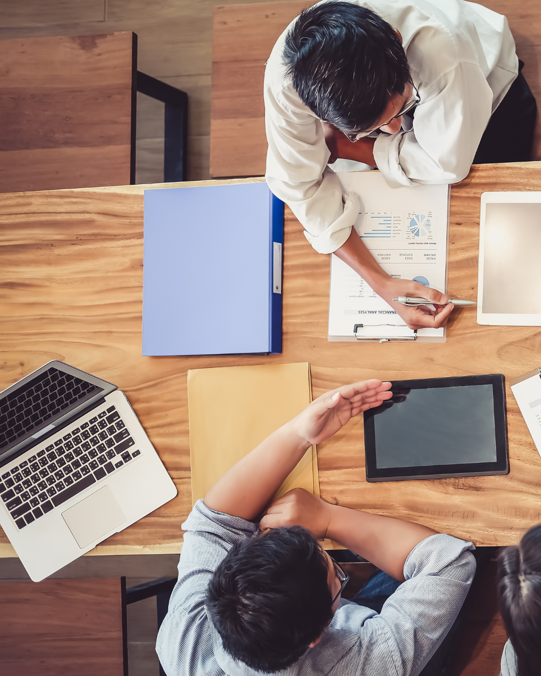 Two people at a wooden table discussing work documents with a binder, laptop, tablet, and clipboard.