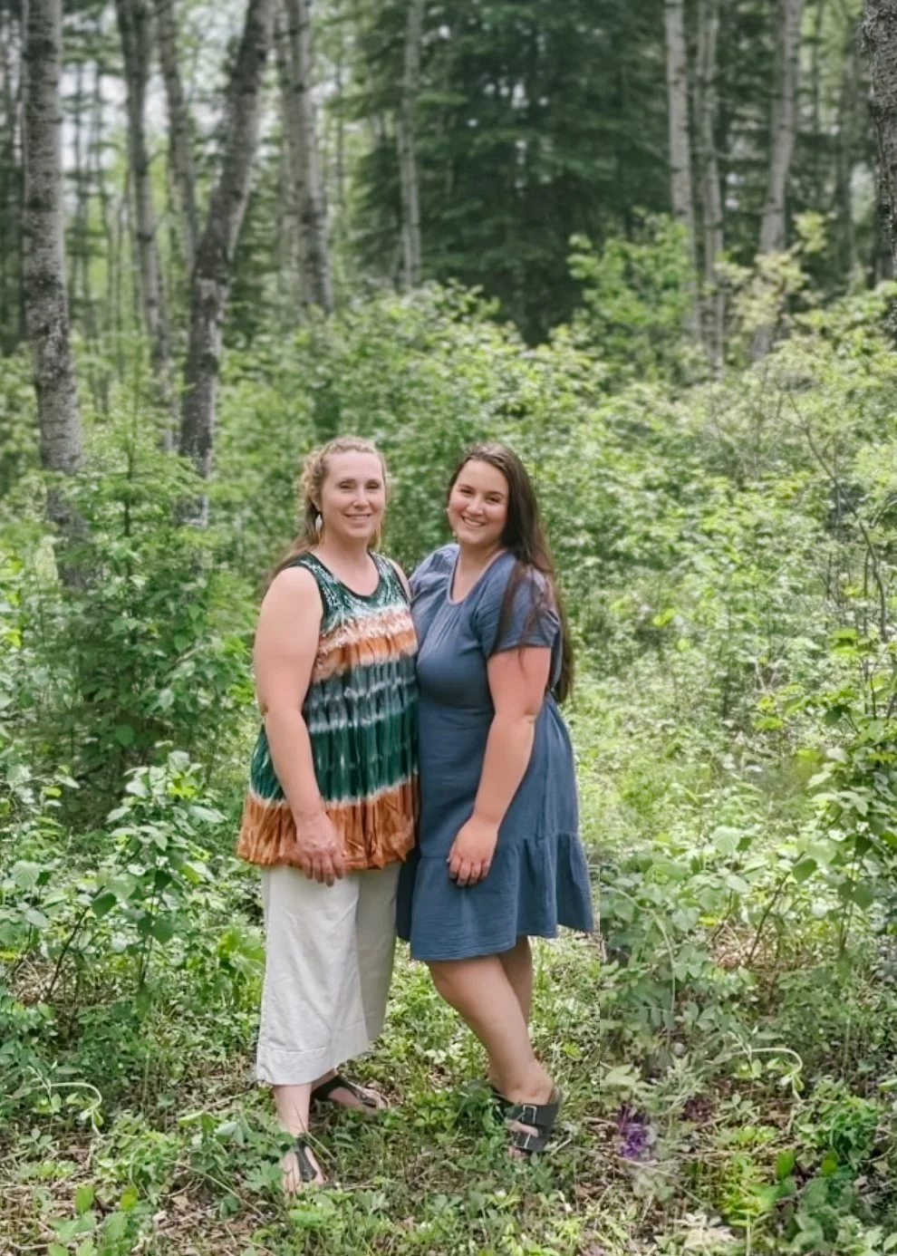 Two women standing together in a lush green forest, smiling at the camera, one wearing a colorful tie-dye dress and the other a blue dress, with trees and foliage in the background.