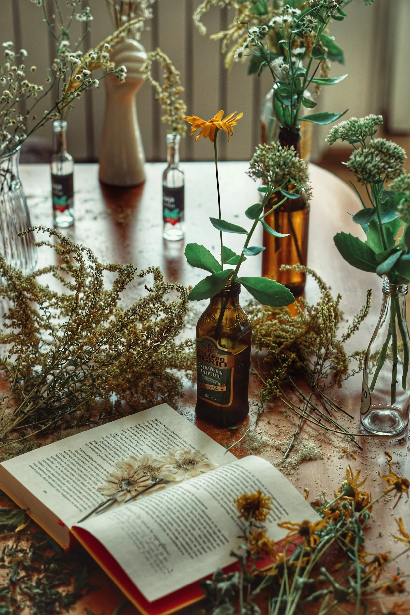 A table decorated with various flowers in glass bottles and vases, an open book with pressed flowers on its pages, and dried flower arrangements surrounding it during a herbal preparation.