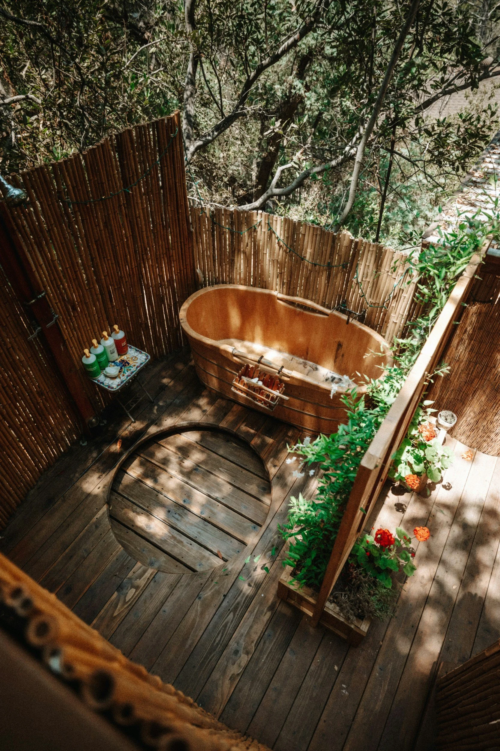 Wooden bathtub on a outdoor deck surrounded by bamboo fencing, with potted plants and sunlight filtering through trees.