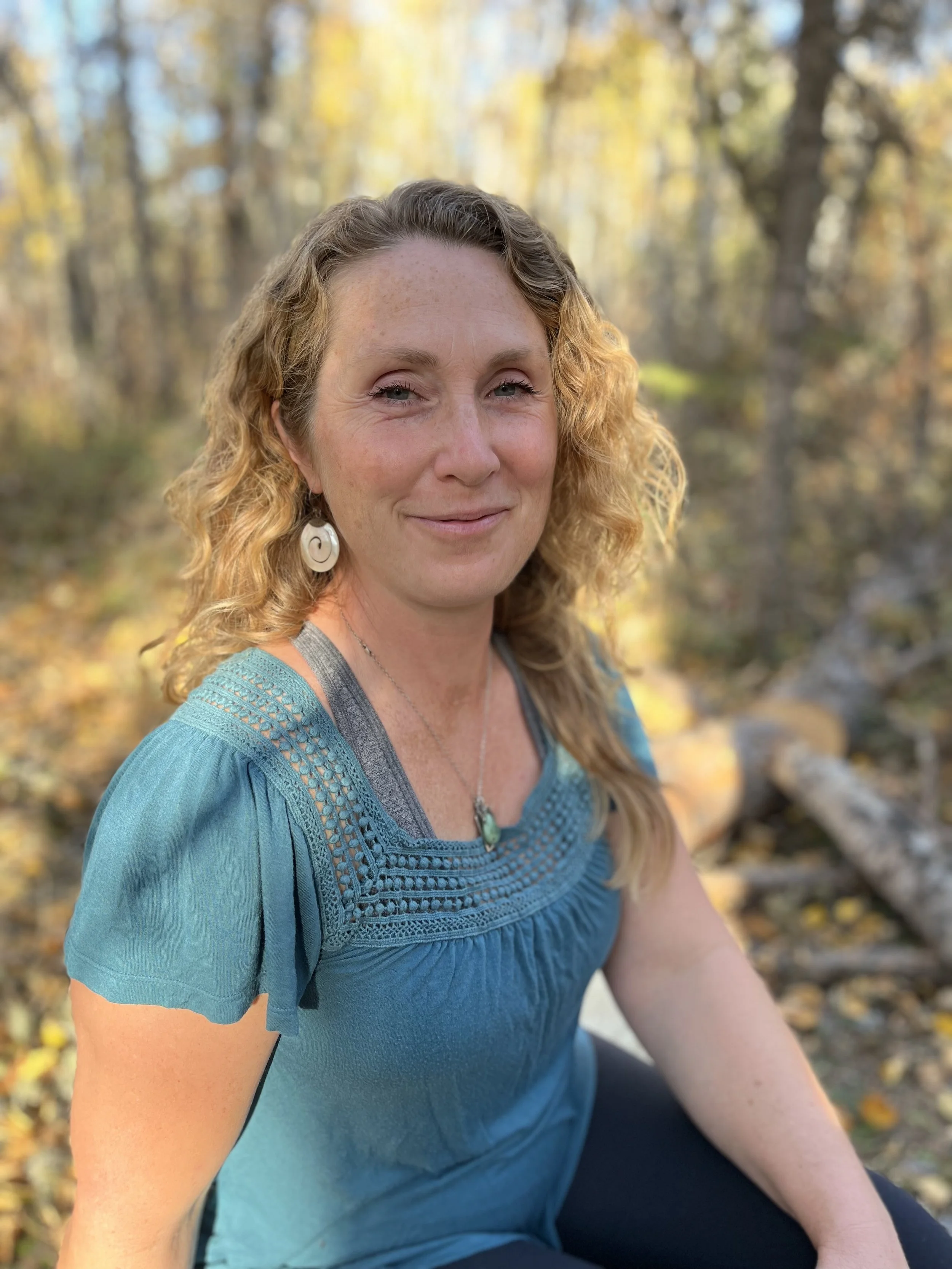 A woman with curly hair wearing a blue embroidered top and jewelry, sitting outdoors in a fall forest with yellow and orange leaves.