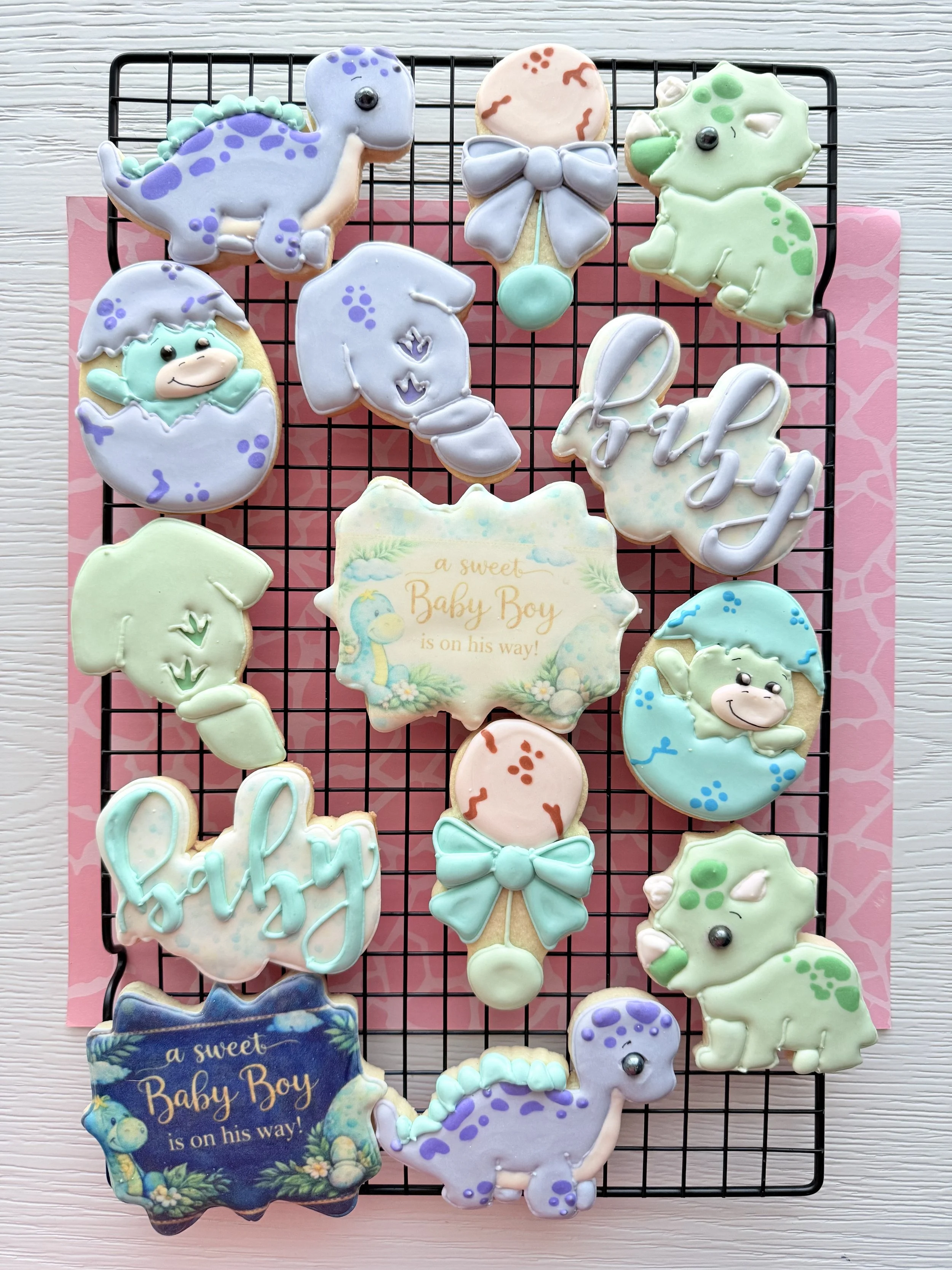 Assorted baby shower cookies on a black cooling rack, featuring dinosaurs, elephants, a sign that says 'a sweet Baby Boy is on his way!', and the word 'baby' in pastel colors.