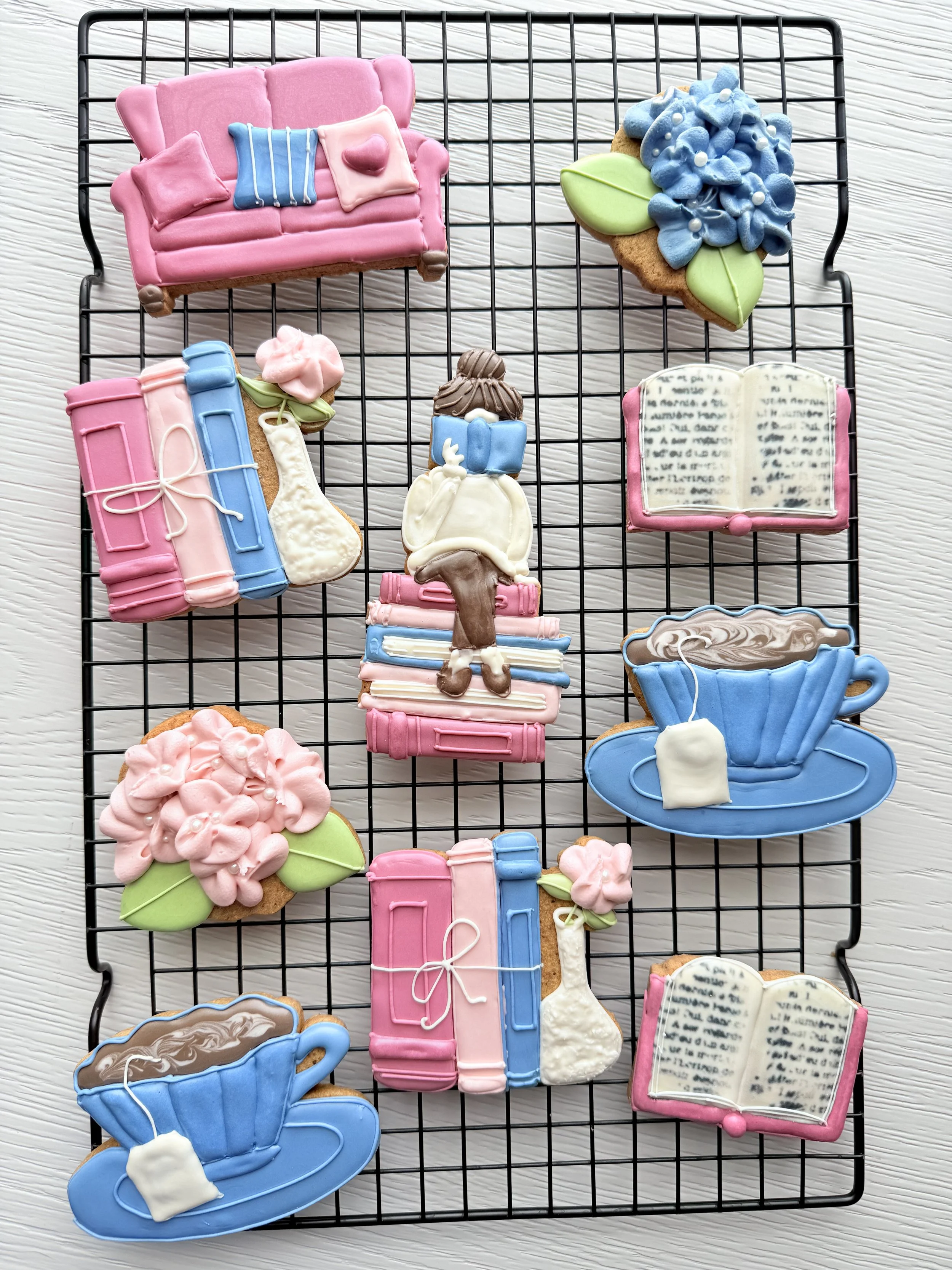Assorted decorated cookies on a black wire cooling rack, including a pink sofa, a blue flower arrangement, a stack of books with a stocking, a girl reading a book, a pink flowers, an open book, and a blue teacup with a saucer, all decorated with colo