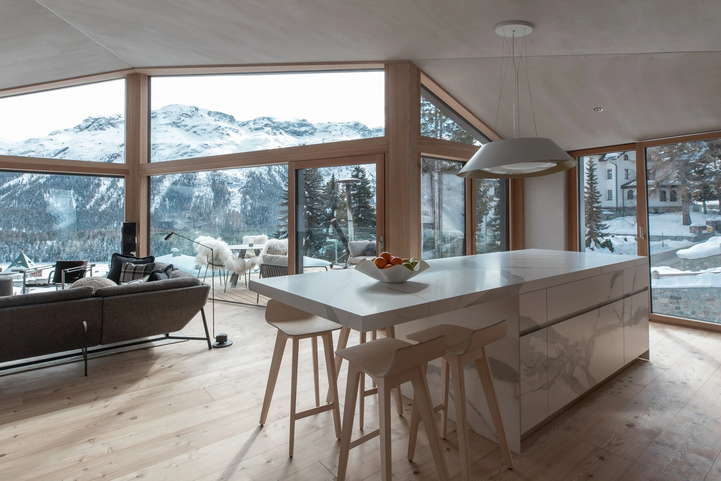Modern open-concept living and kitchen area with large floor-to-ceiling windows showing snowy mountains and a perfect view over St. Moritz and its lake.