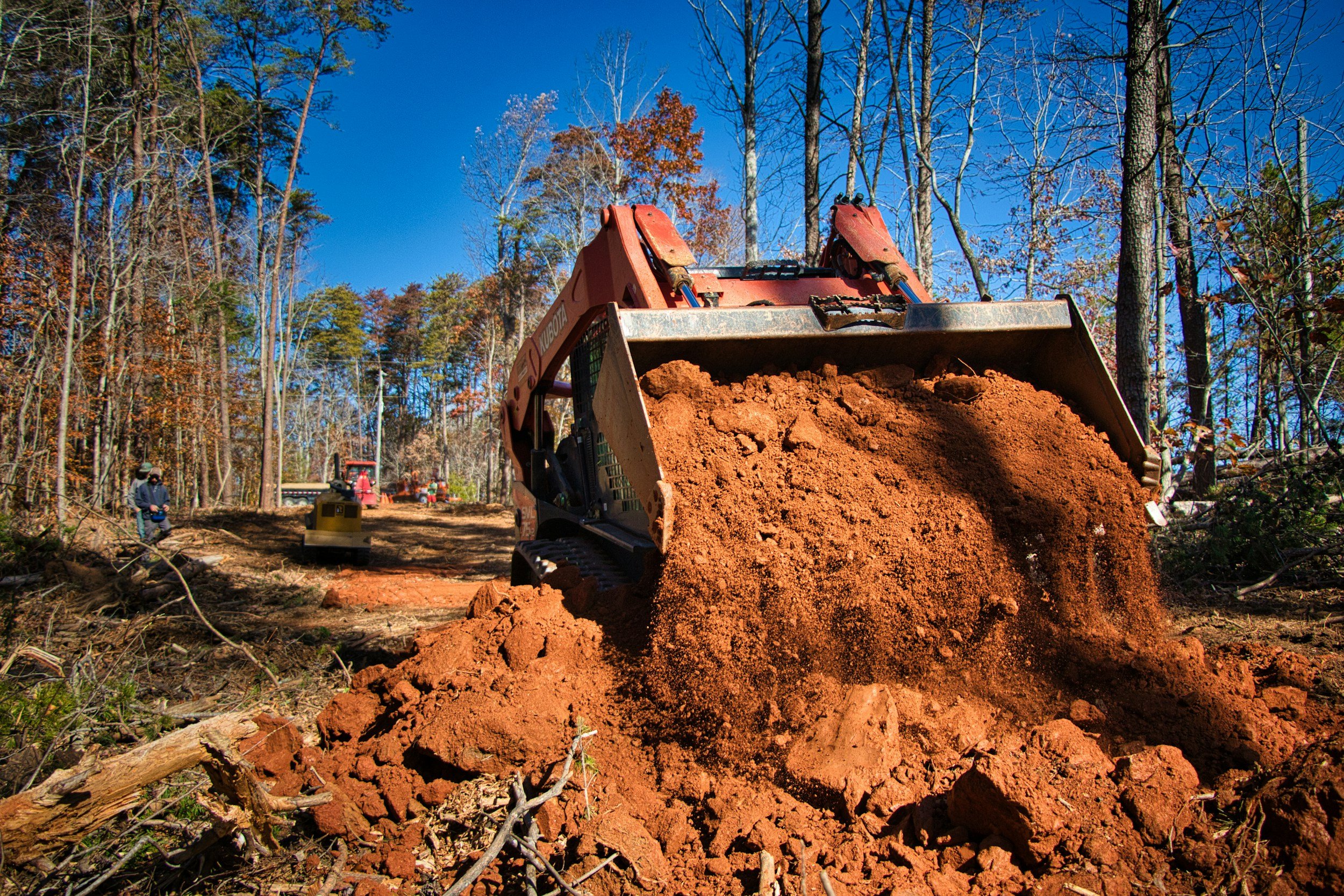 A construction site in a forest with a red excavator moving dirt, workers in gloves and safety gear, and trees with autumn foliage under a clear blue sky.