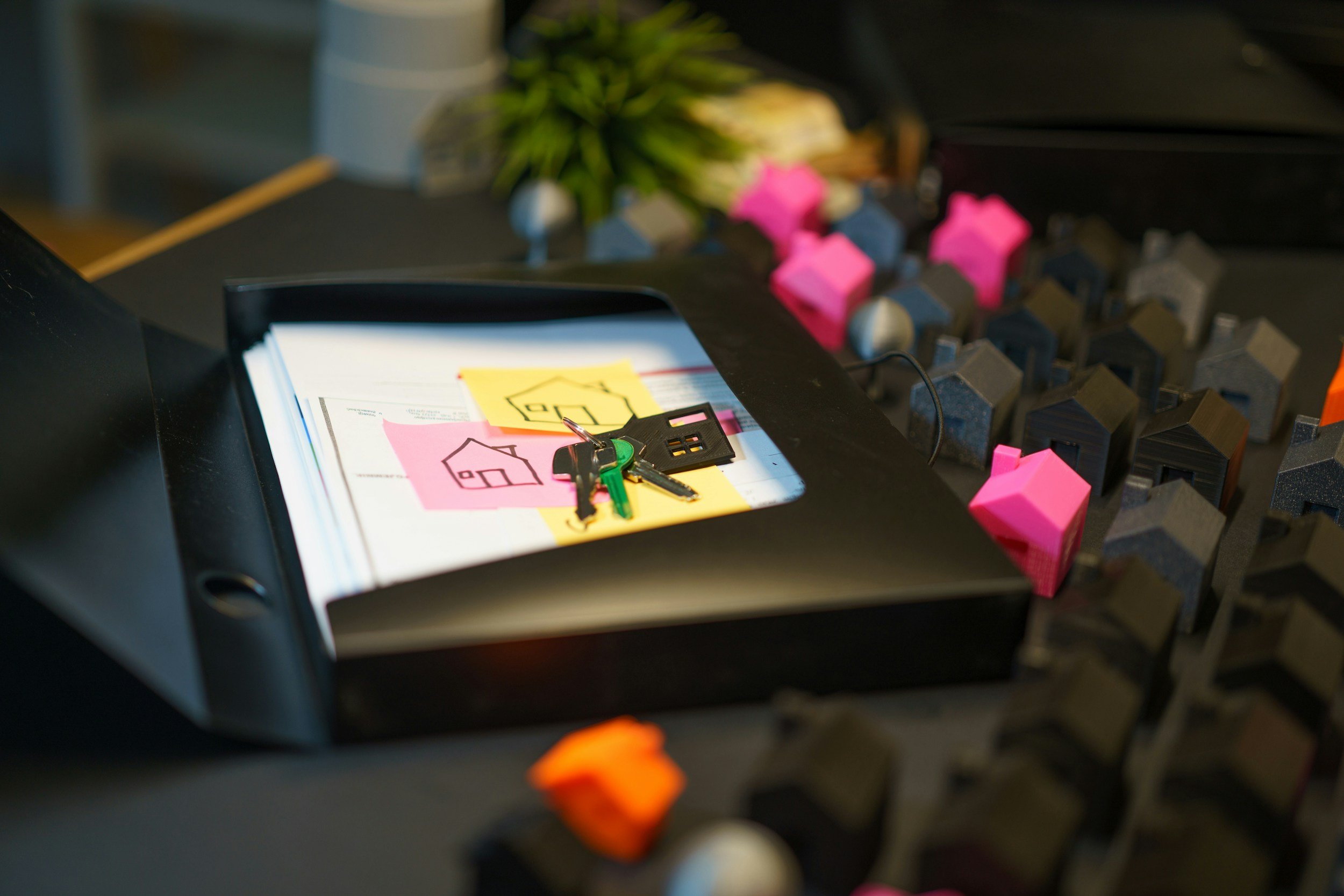 A desk with a black binder containing papers, keys on top, and keys on a keychain, with various colored miniature house models surrounding it.
