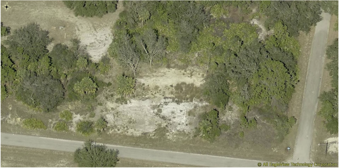 Aerial view of a semi-wooded area with a dirt patch and surrounding trees, next to a paved road.