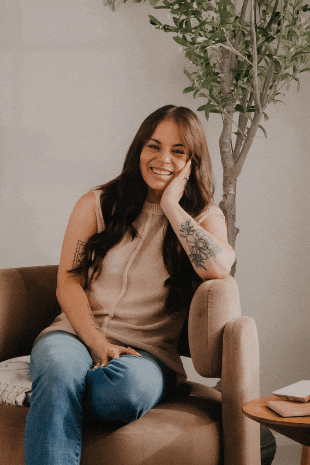 A woman with brown, wavy hair sitting on a beige armchair, smiling, with a tattoo of flowers on her arm, wearing a sleeveless beige top and blue jeans, seated indoors with a potted plant and books nearby.