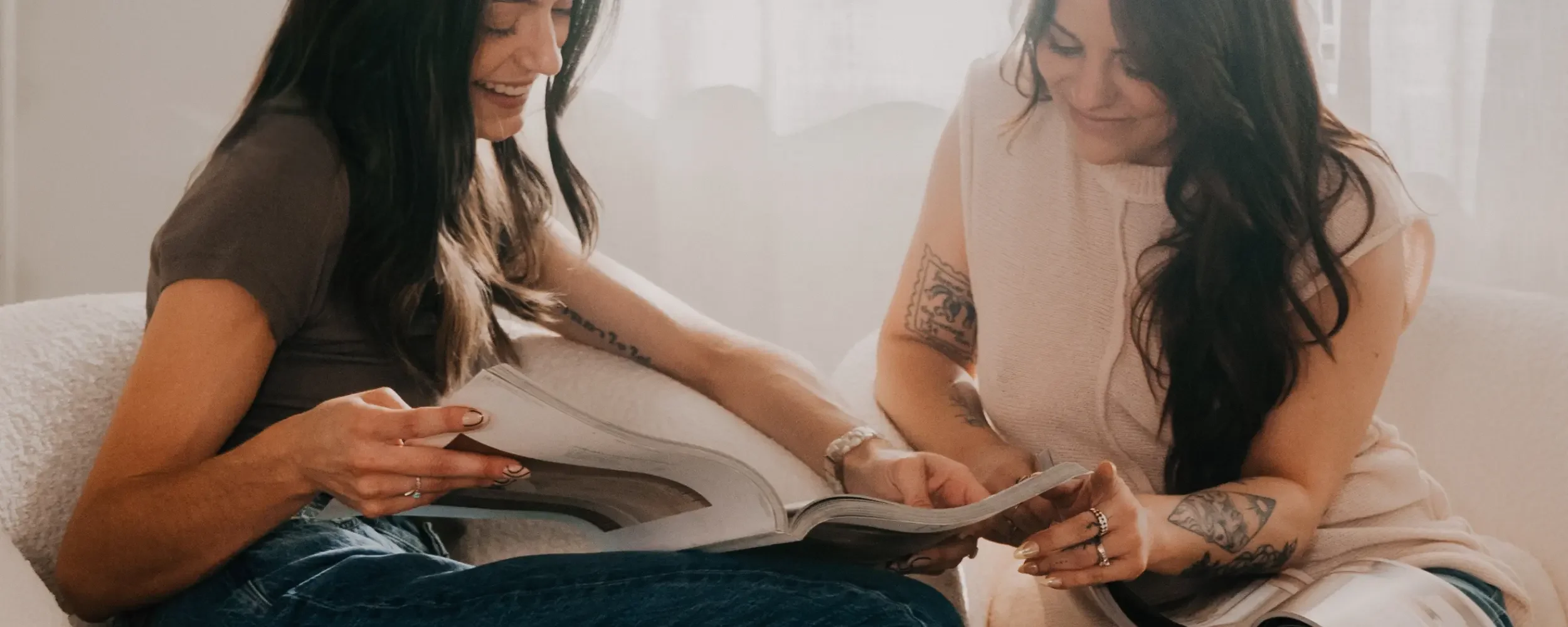 Two women sitting on a couch, looking at a magazine together, smiling and sharing a moment of laughter.