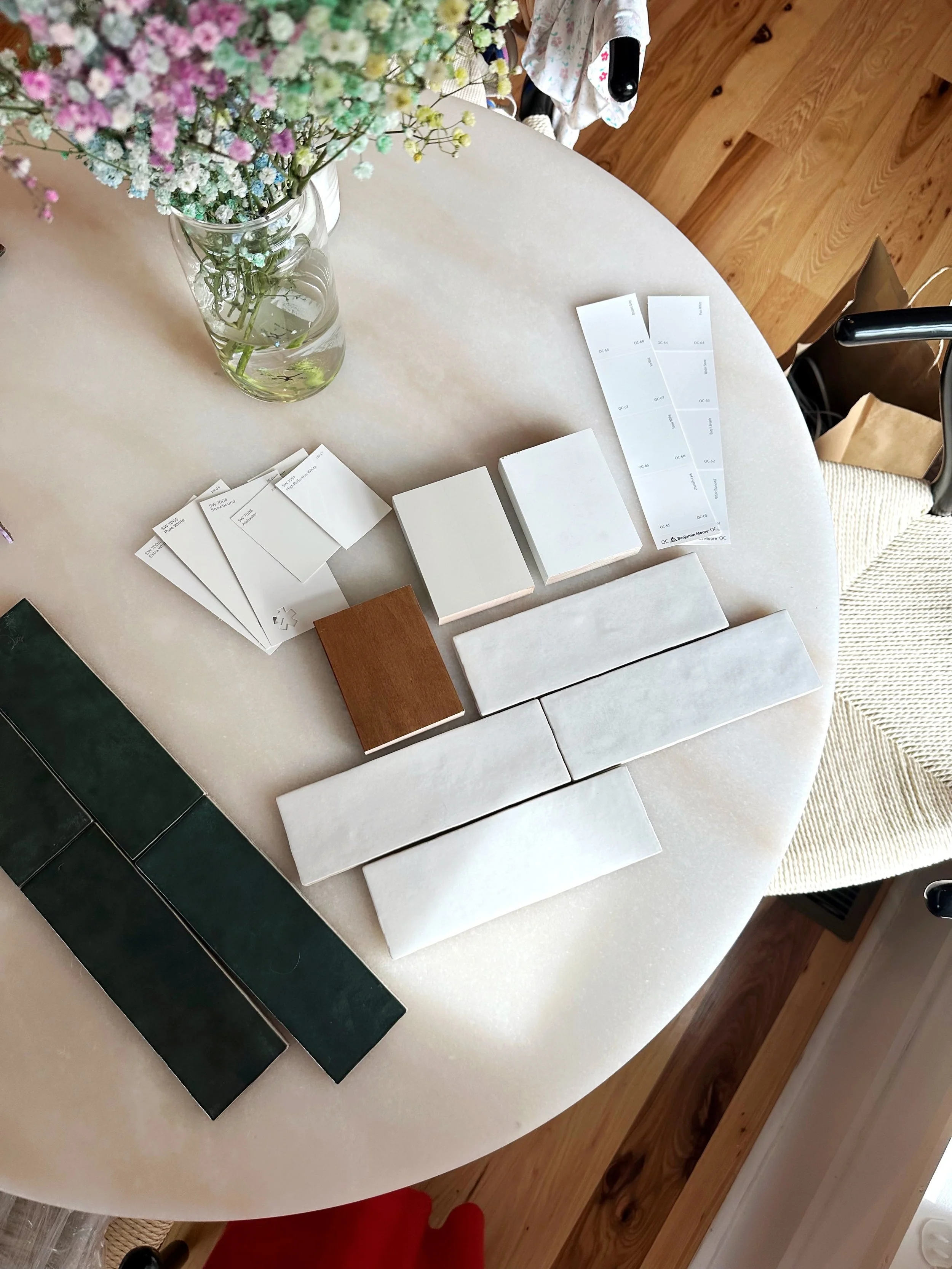 A round table with a vase of multicolored flowers, various white tiles, a small brown tile sample, brochures, and samples of green tiles.