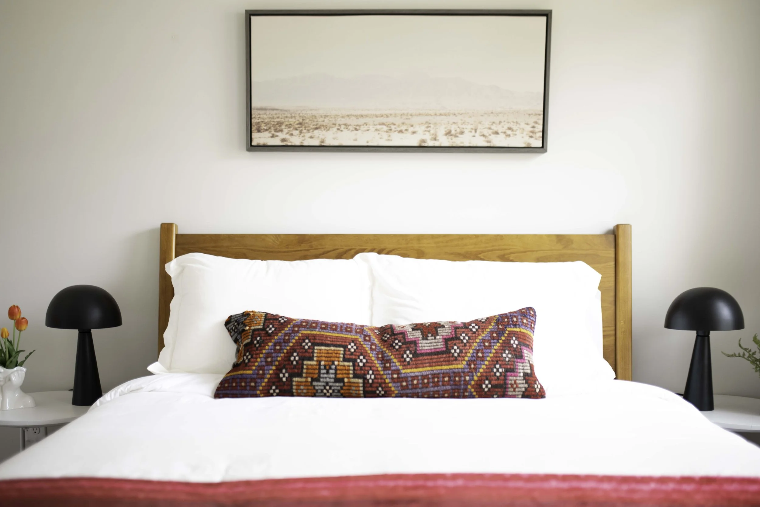 A neatly made bed with white linens and a colorful patterned lumbar pillow, flanked by black table lamps on white bedside tables, in a room with a minimalist landscape painting above the wooden headboard.
