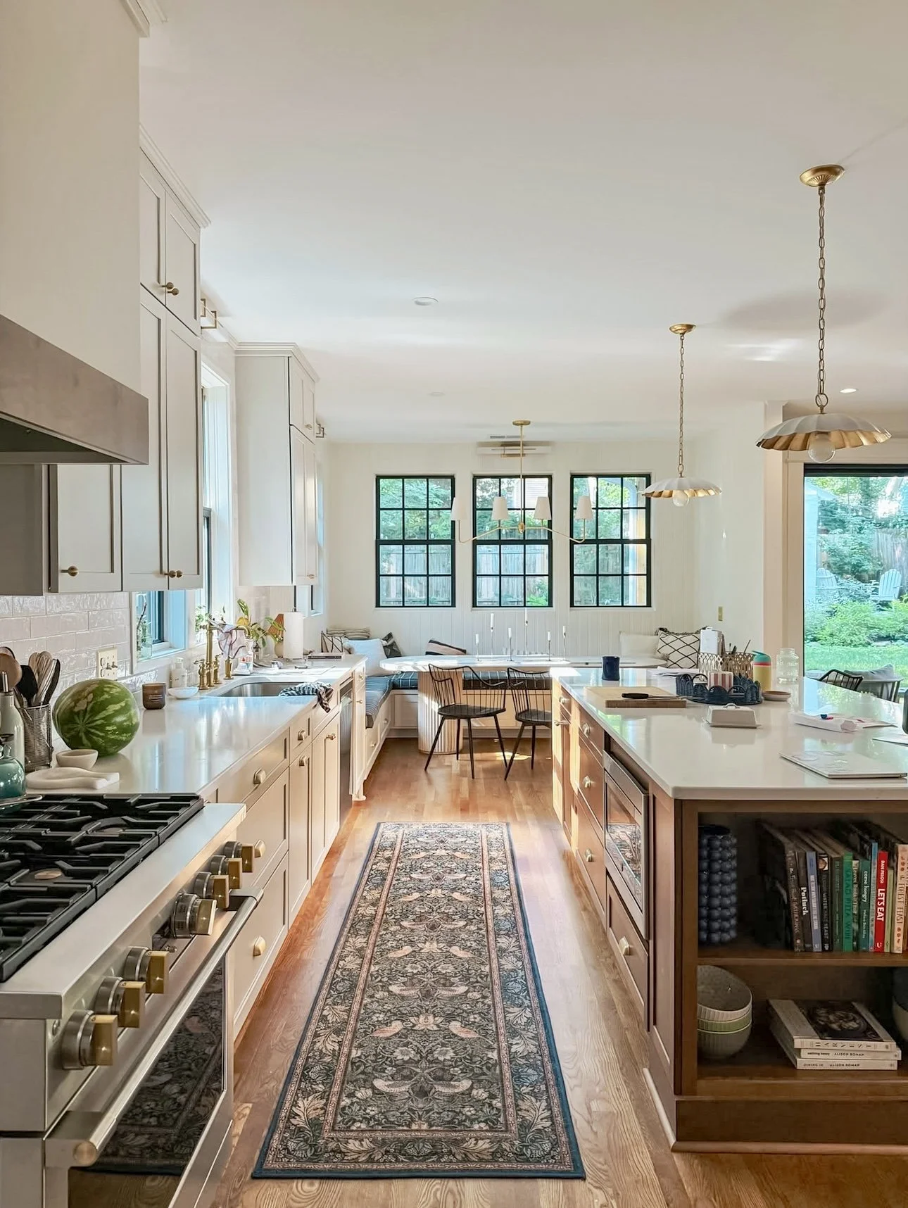 Bright kitchen with white cabinetry, wooden countertops, and a floral patterned runner rug on hardwood floor. Features a stove, bookshelves, a watermelond, and a sitting nook with windows at the back.