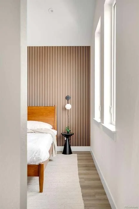 A minimalist bedroom with a wooden bed frame, white bedding, a black side table with a small plant, and a decorative wooden slat accent wall. Natural light streams in from two tall windows.