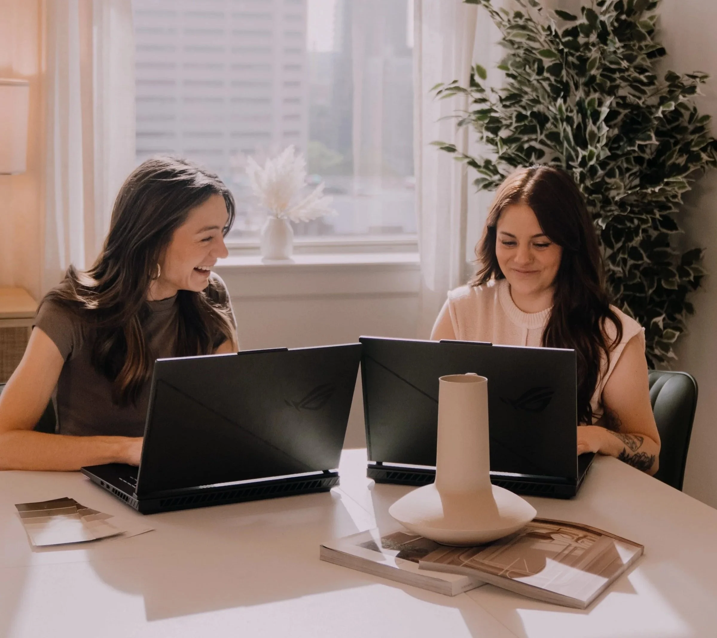 Two women sitting at a table with laptops, smiling and chatting, in a bright room with large windows, a tall green plant, and decorative items on the table.