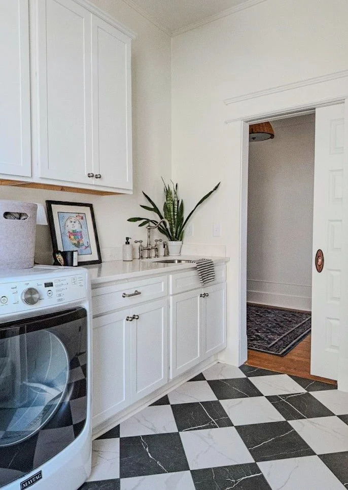 Laundry room with white cabinets, a washer, a framed colorful drawing, a potted plant, soap dispenser, striped dish towel, and patterned black and white tile floor