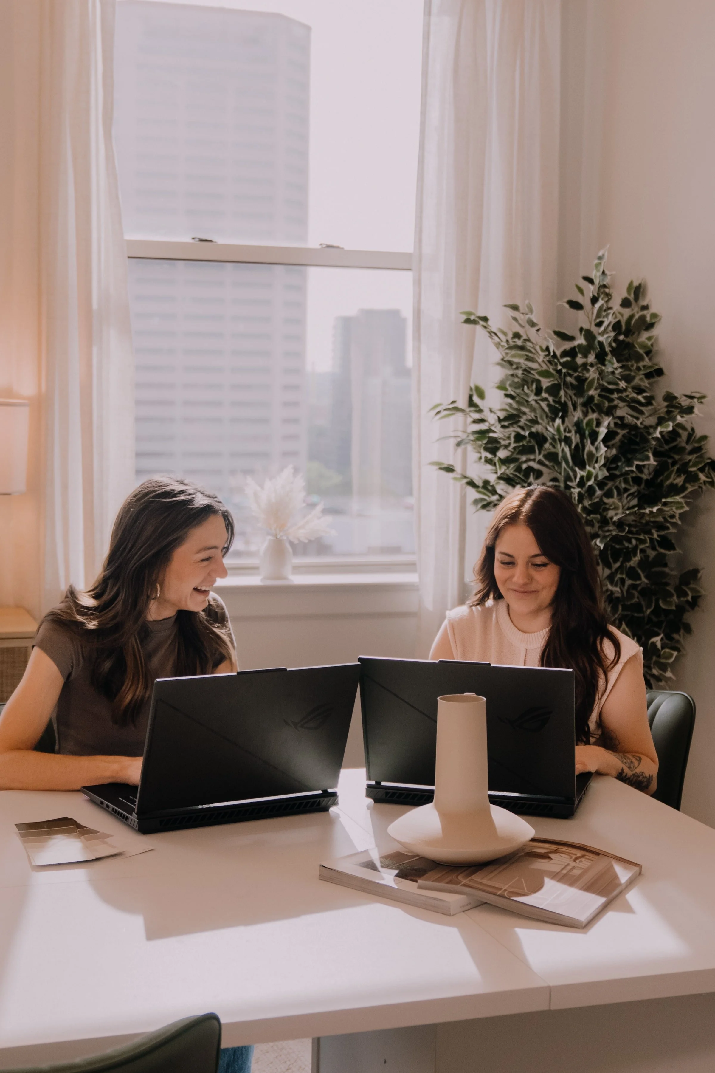 Two women sitting at a white table, working on laptops, and smiling in a sunlit room with large window, potted plant, and decorative items.