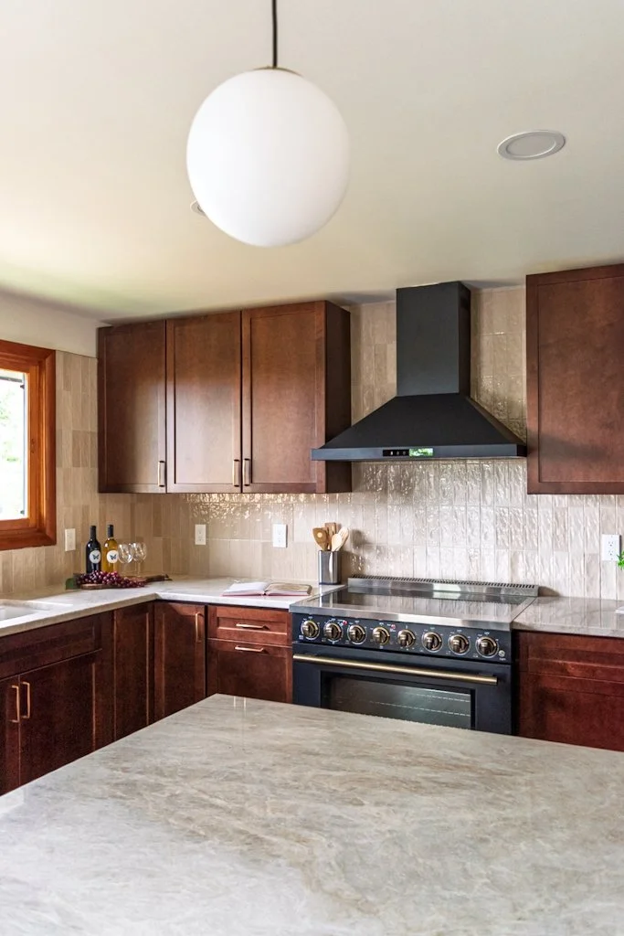 Modern kitchen with dark wood cabinets, beige tiled backsplash, black range hood, stainless steel stove, and a large island with a marble countertop