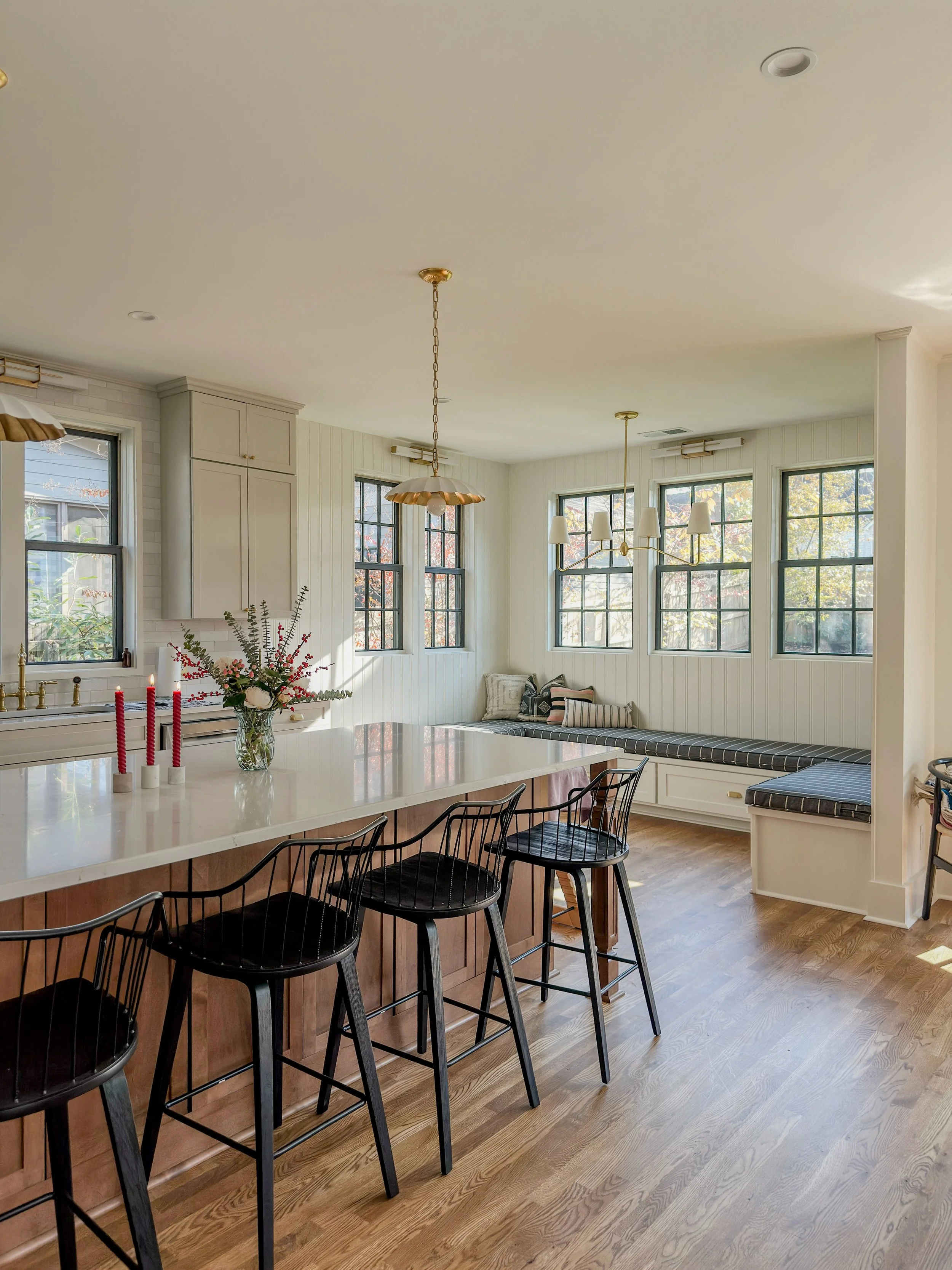 Bright kitchen with white cabinets, large island with bar stools, window seating with cushions, hardwood floors, and multiple windows letting in natural light.