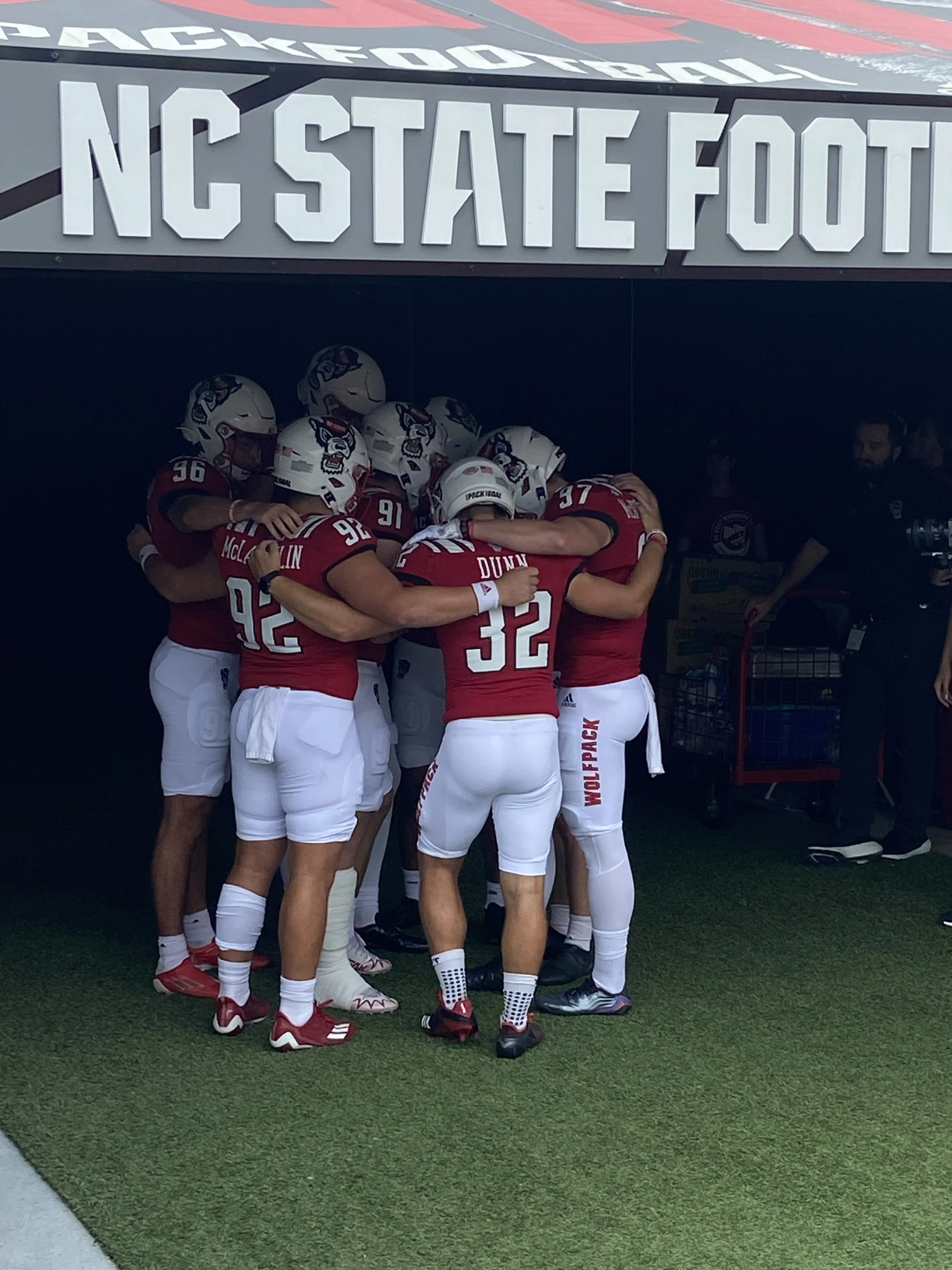American football players in red jerseys and white pants huddling together under a sign that reads 'NC State Football' at a football stadium.