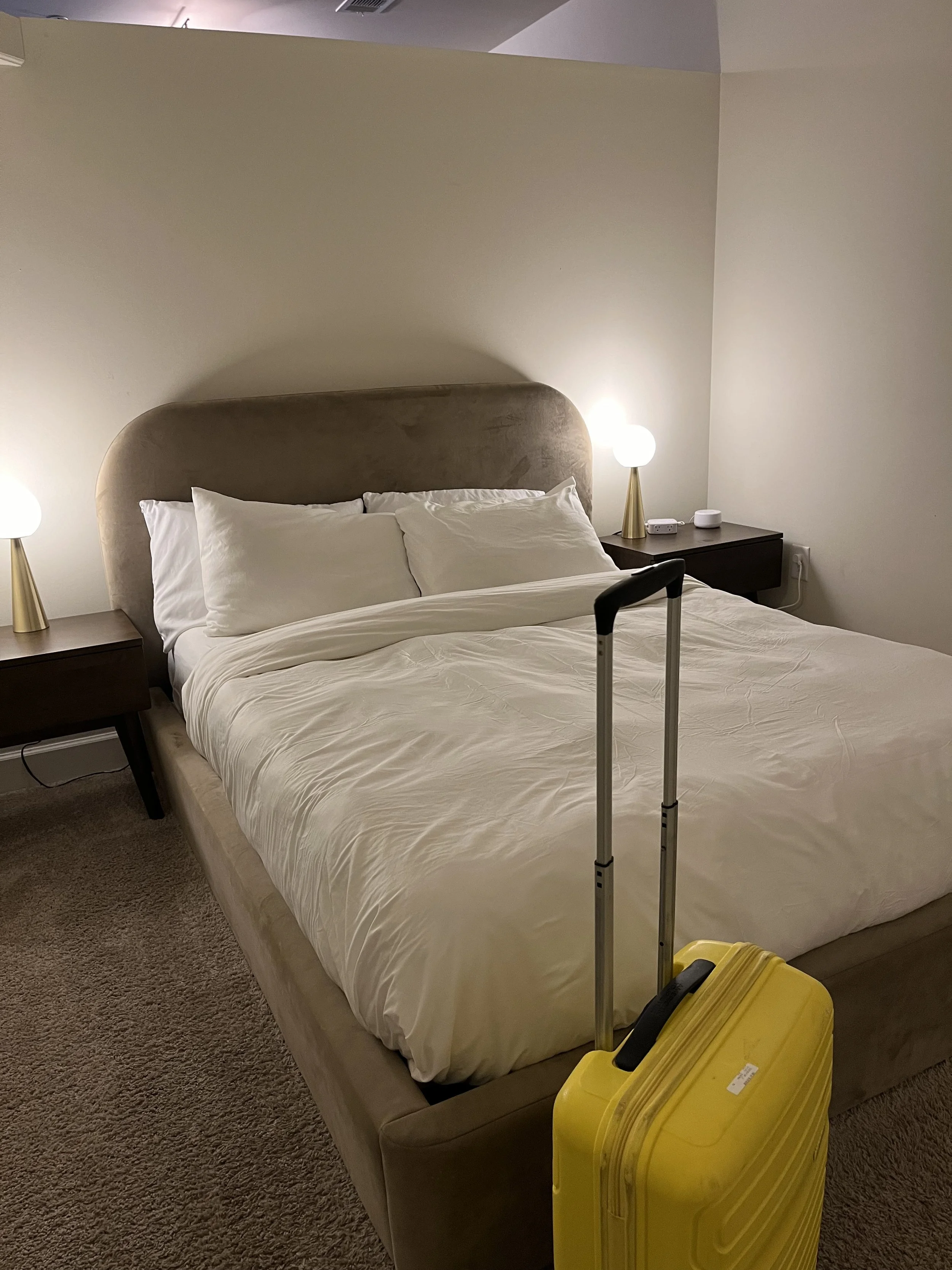 A hotel room with a bed, bedside tables, and lamps, with a yellow suitcase with a black handle and extended handle in front of the bed.