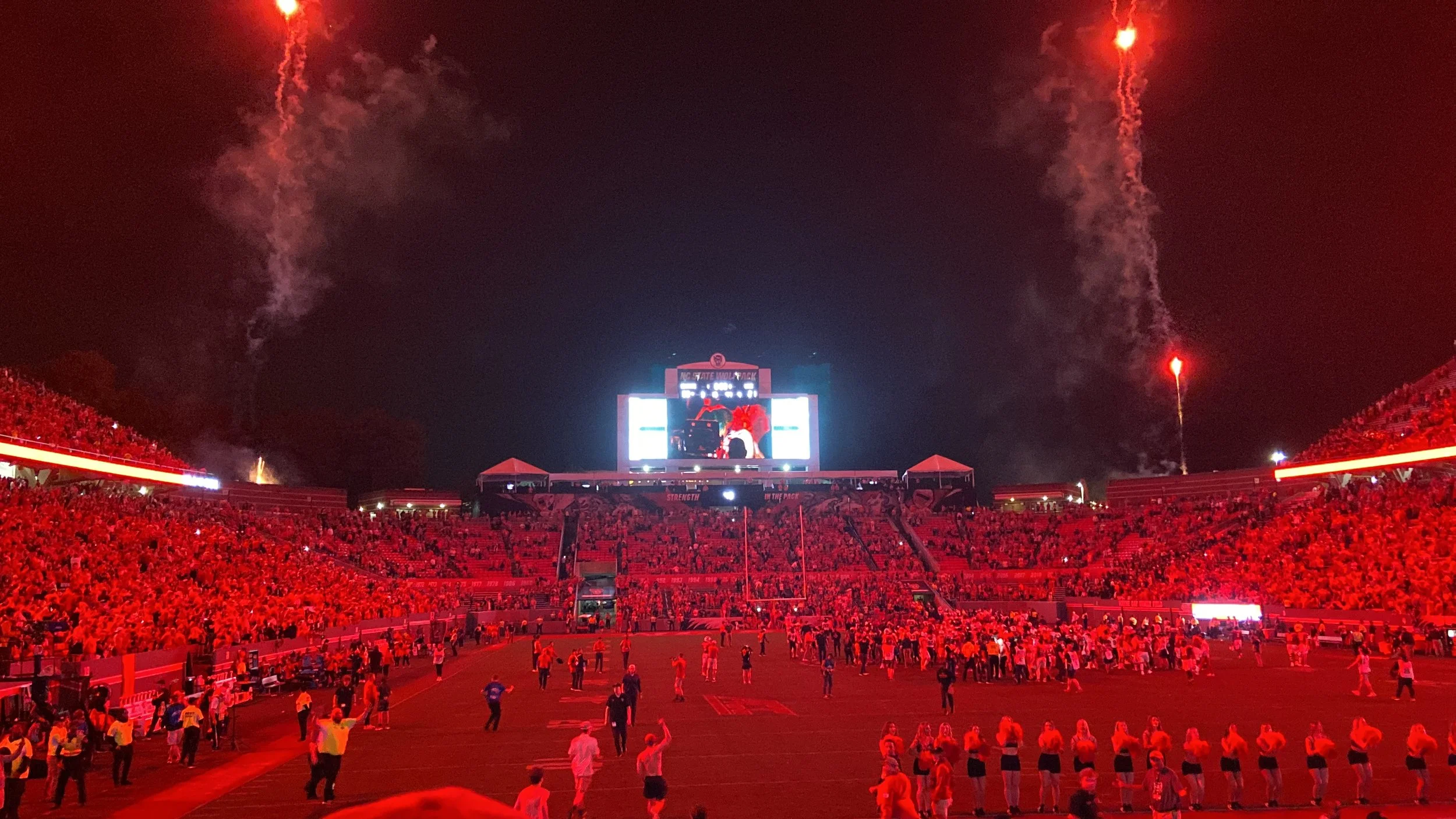 The Carter-Finley Stadium filled with fans at night, with red lighting, fireworks in the sky, and a large screen displaying a live game or event.