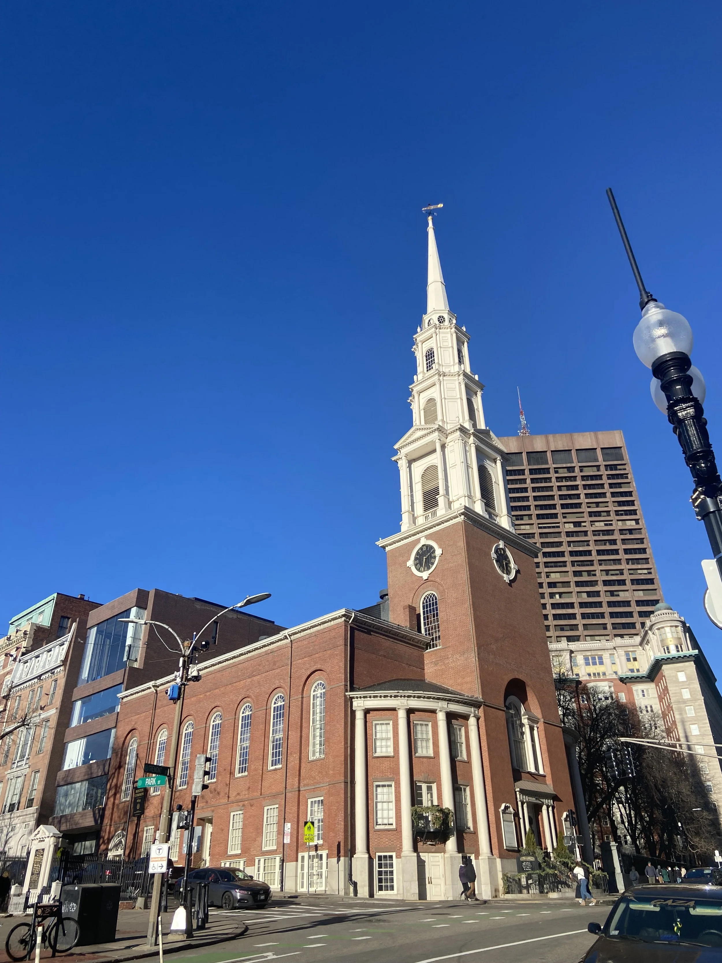 A historic brick church building with a tall white steeple and clock faces, set against a bright blue sky in an urban area.