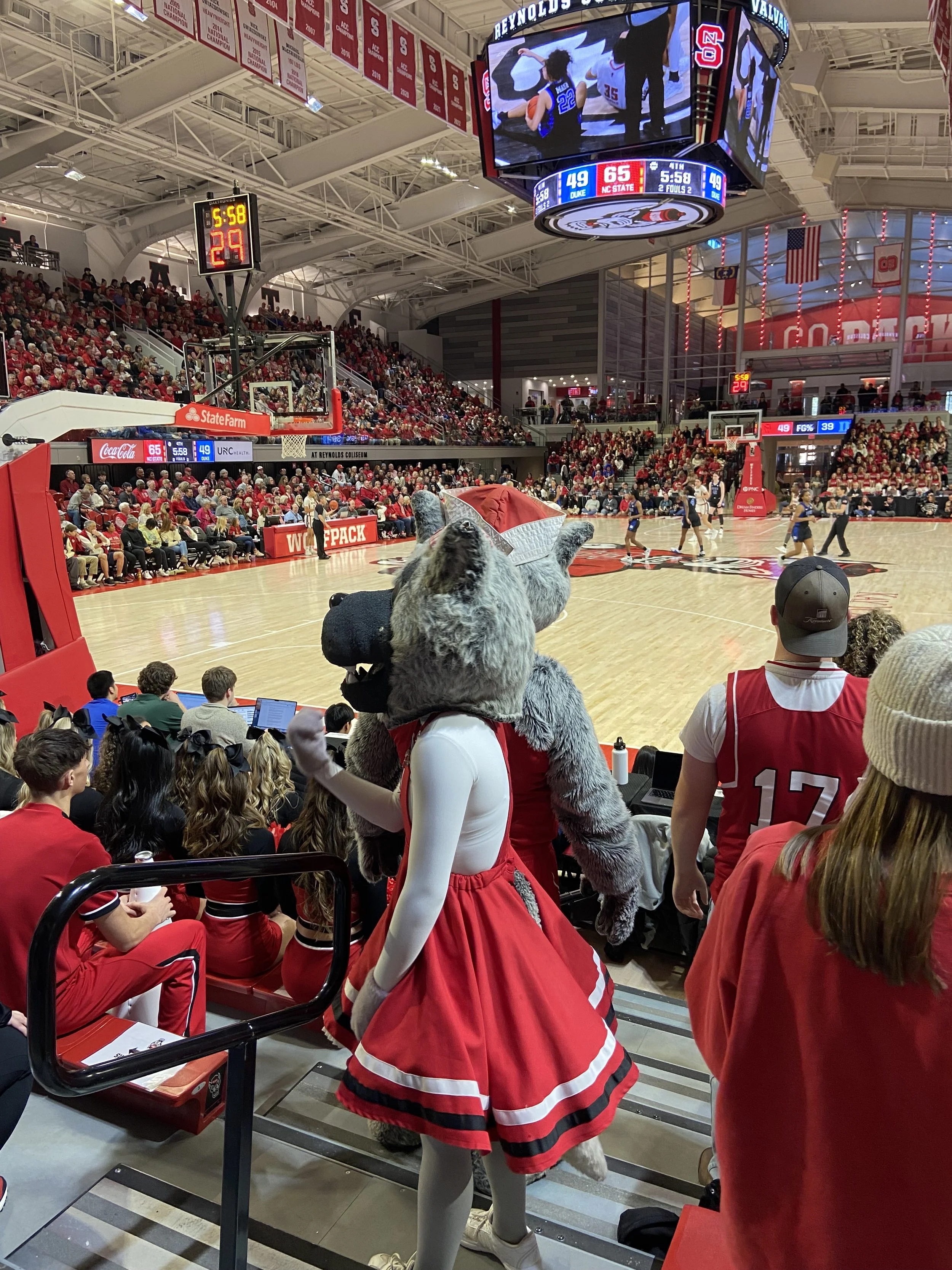 A person dressed in a wolf mascot costume and a cheerleader in red dress at a basketball game in an indoor arena, with many spectators in the stands.