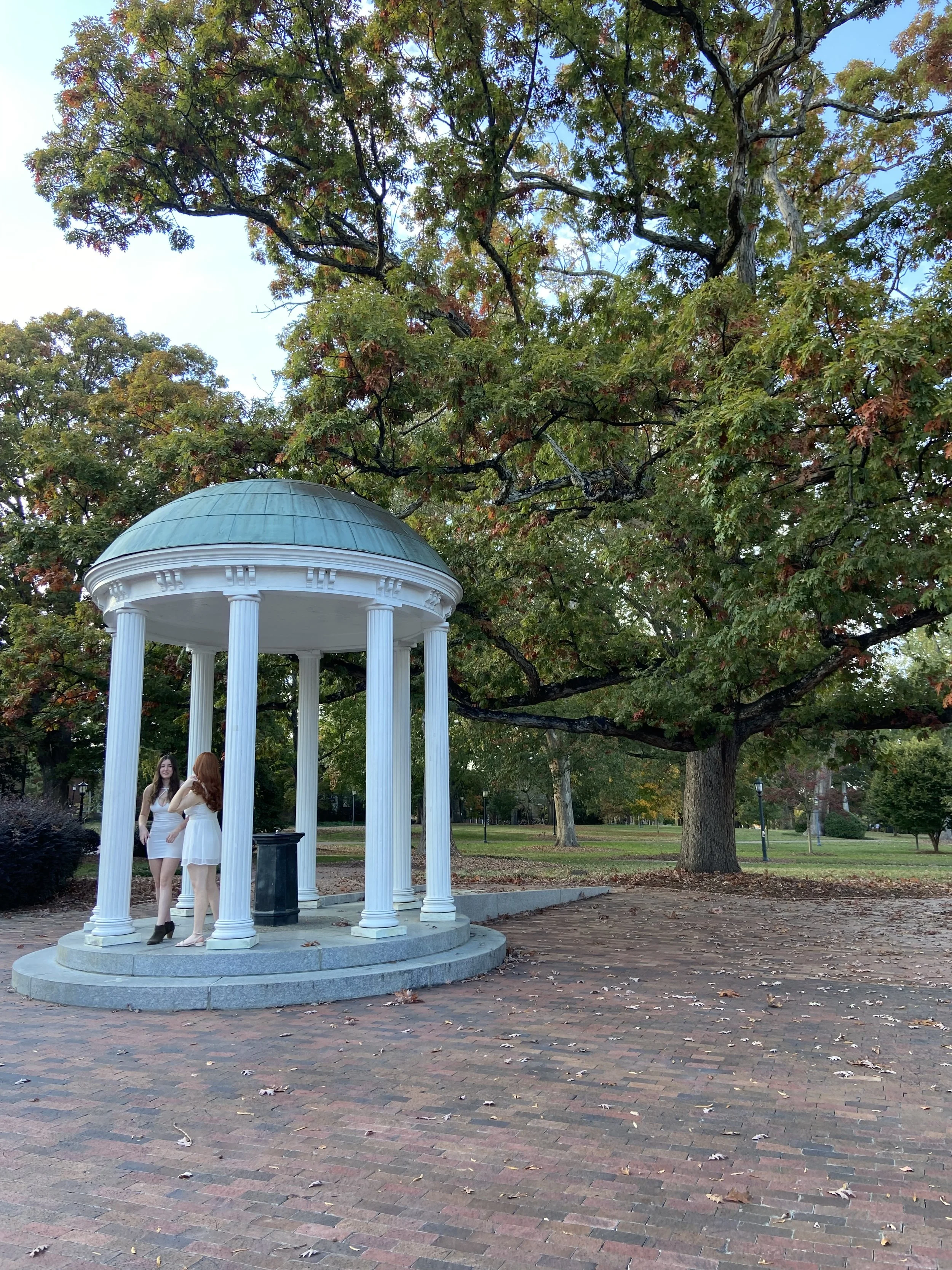 The small, white, neoclassical pavilion at UNC with columns and a domed roof in a park, with three women dressed in white standing underneath, near a large tree with changing leaves.
