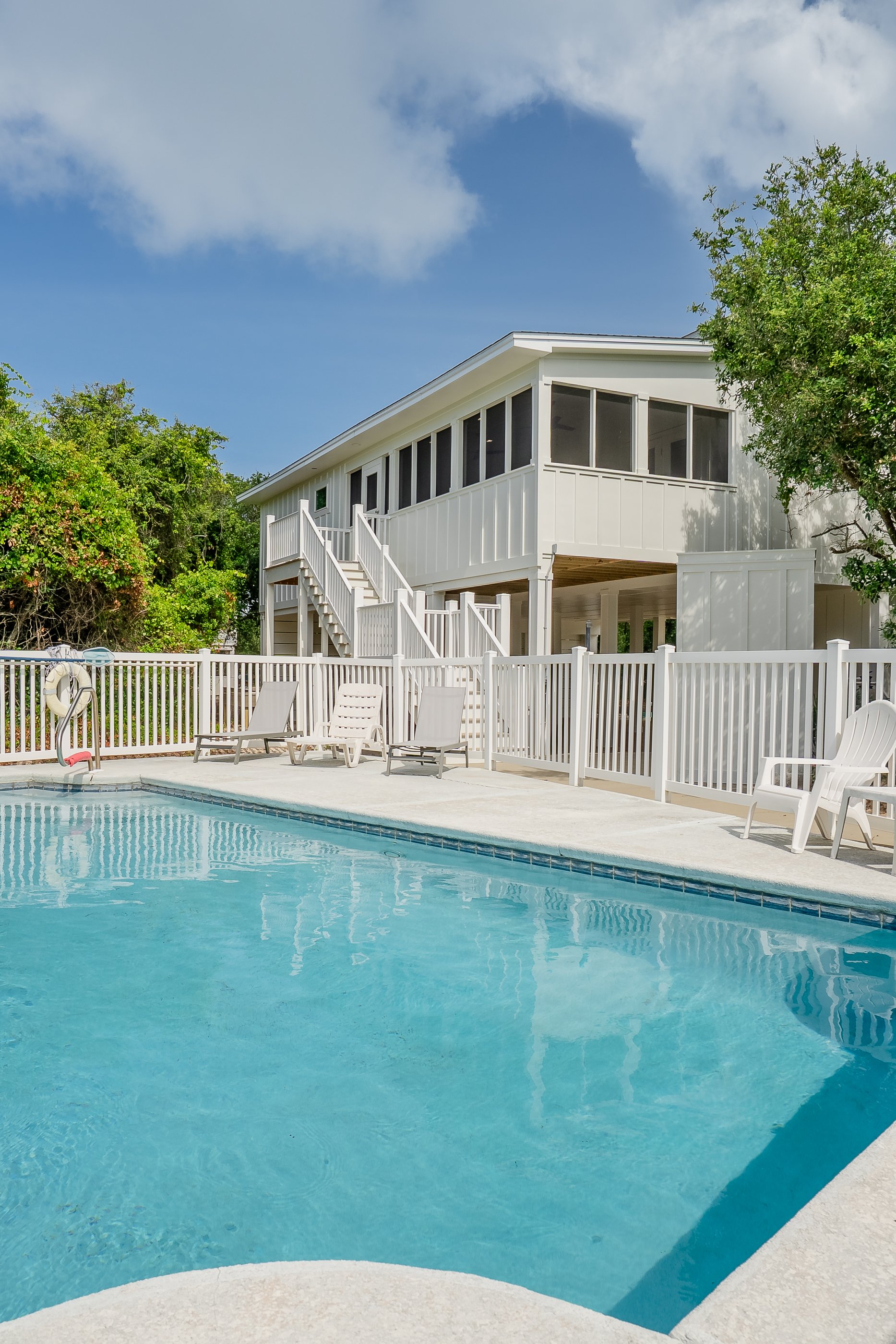 A backyard with a swimming pool, white lounge chairs, and a raised house with a staircase, surrounded by green trees on a sunny day.