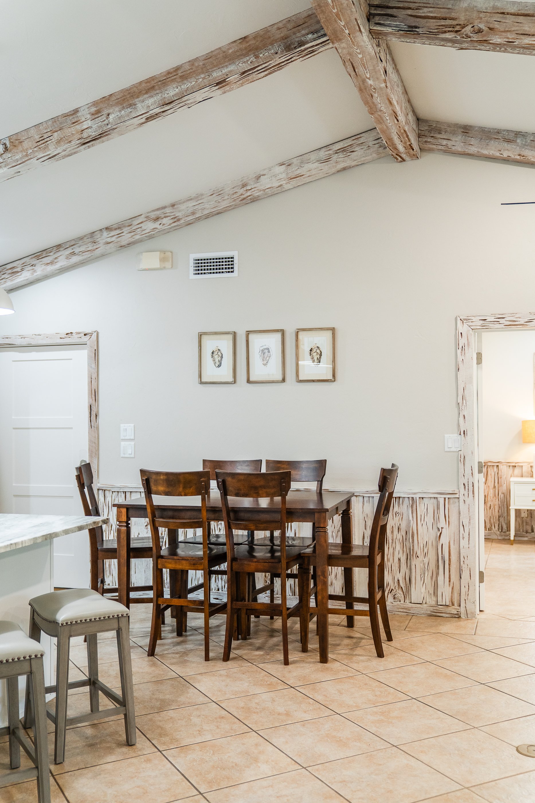 Dining area with a wooden table and six matching chairs in a room with a pitched ceiling and rustic wood trim. There are three framed pictures on the wall and a doorway leading to another room.