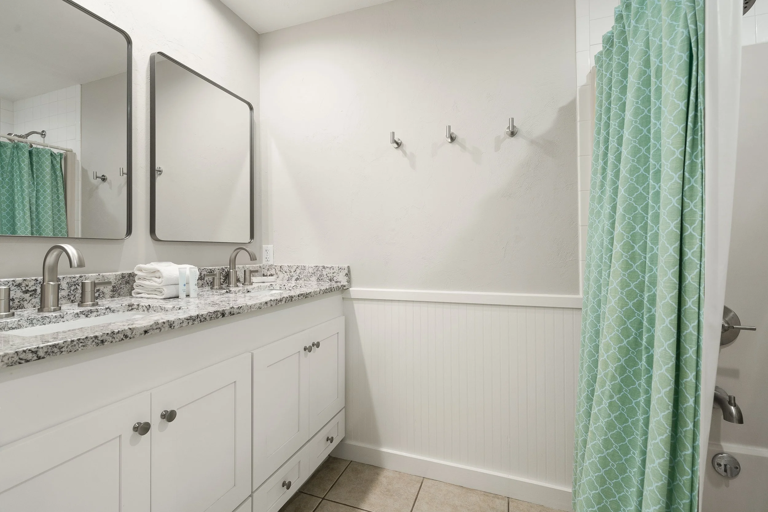 Bathroom with dual sinks, granite countertops, two mirrors, white cabinets, towel, hooks on the wall, and turquoise shower curtain.