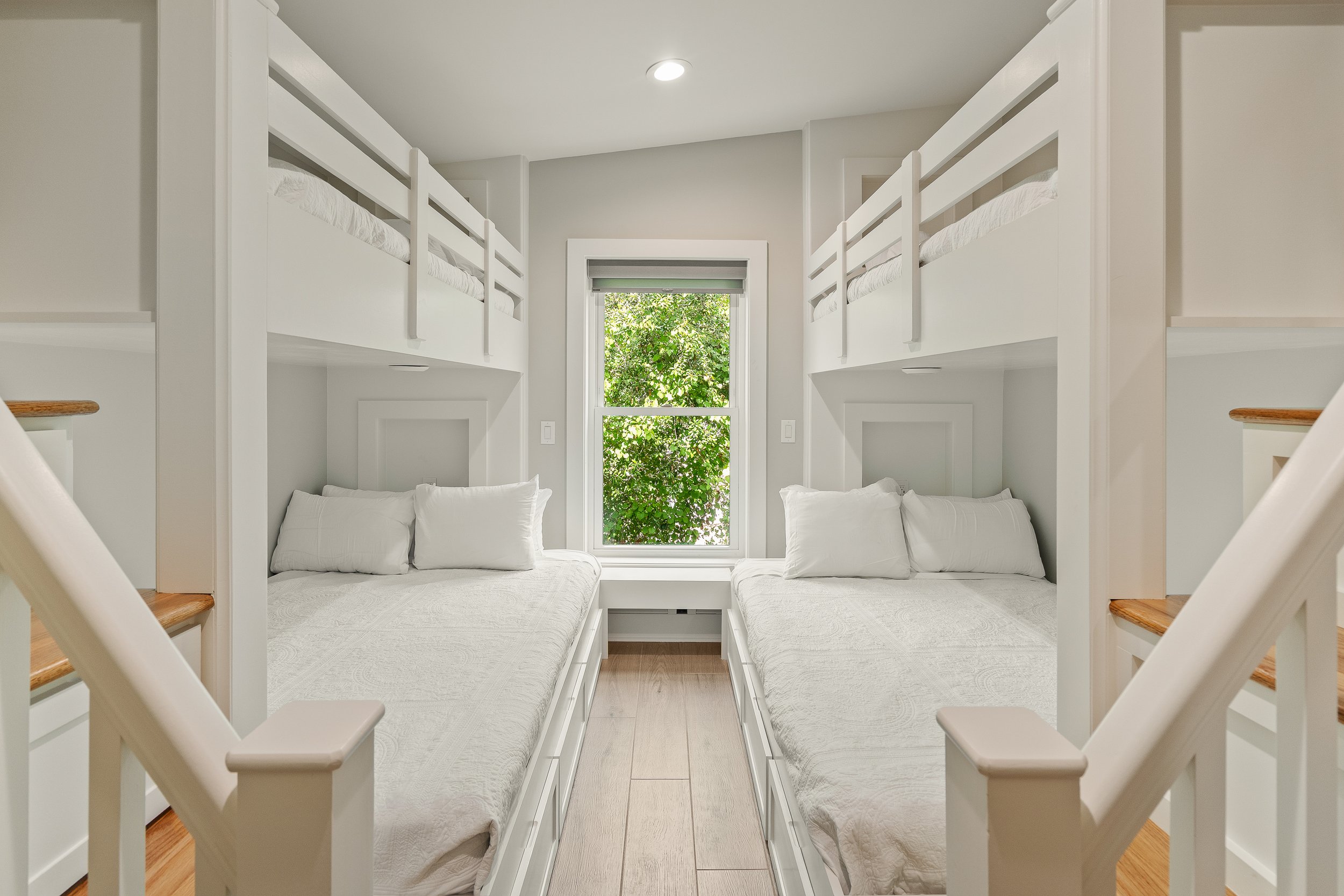 Two sets of twin beds with white bedding, built-in storage drawers underneath, and a central window with green foliage outside, in a small, bright bedroom.