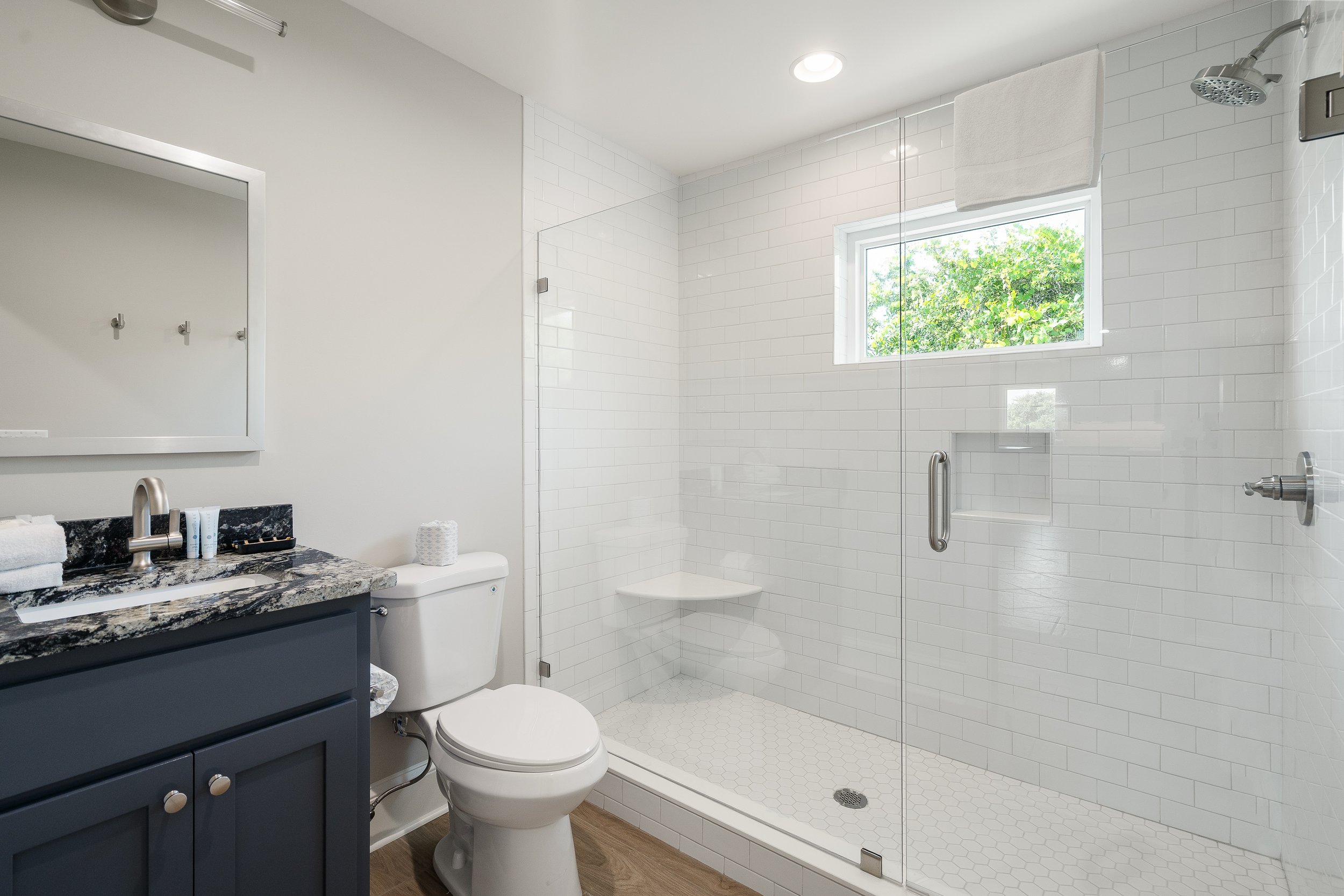 Modern bathroom with walk-in shower, white subway tiles, small window, blue vanity with granite countertop, toilet, and wall mirror.