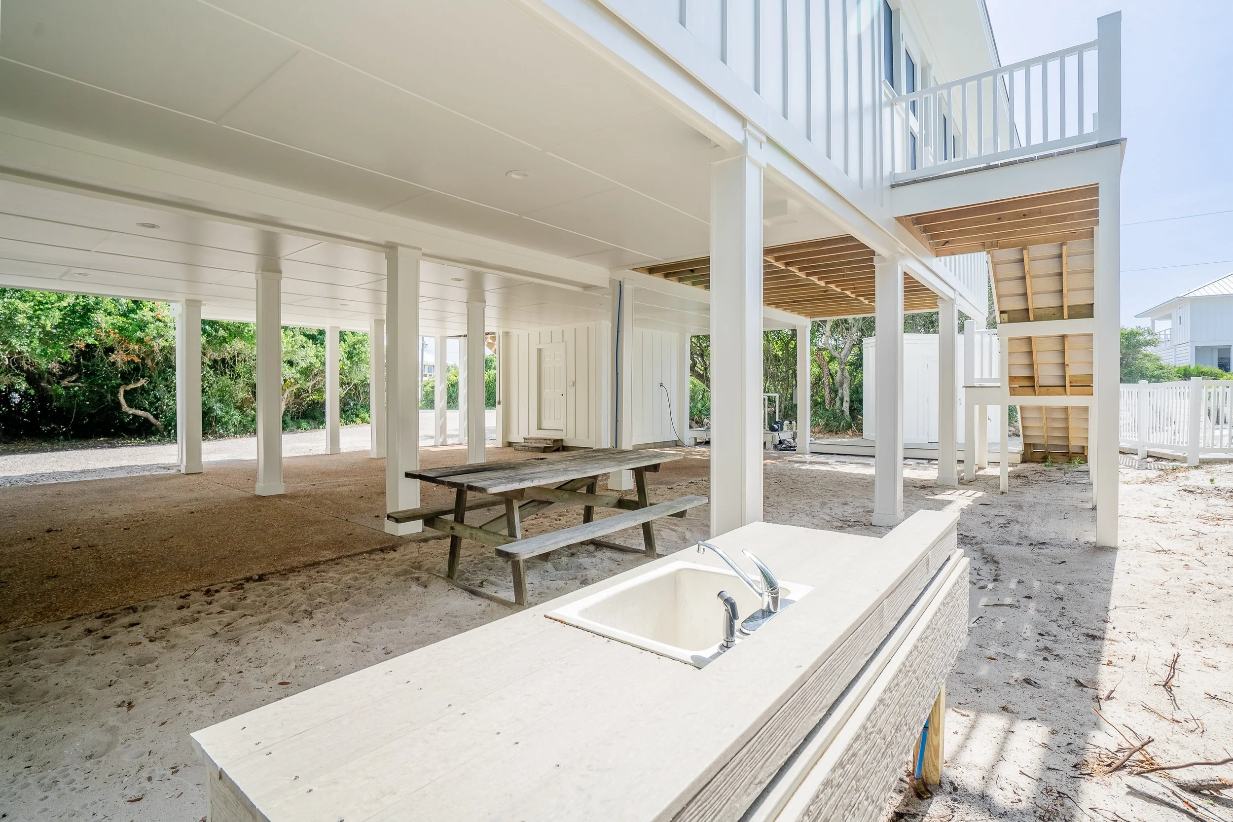 Under-construction elevated house with white support beams, sandy ground, and a built-in outdoor sink, with trees and neighboring houses in the background.