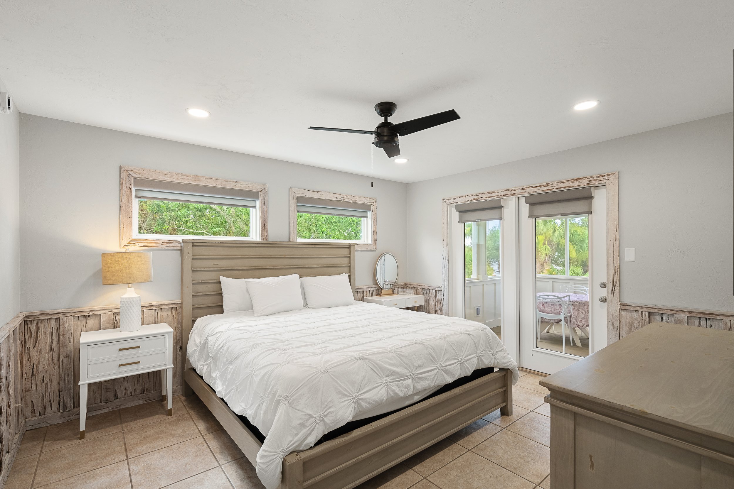 Bedroom with a large bed, nightstand with lamp, small vanity, and balcony with outdoor seating, decorated in neutral tones with wood accents and small horizontal windows.